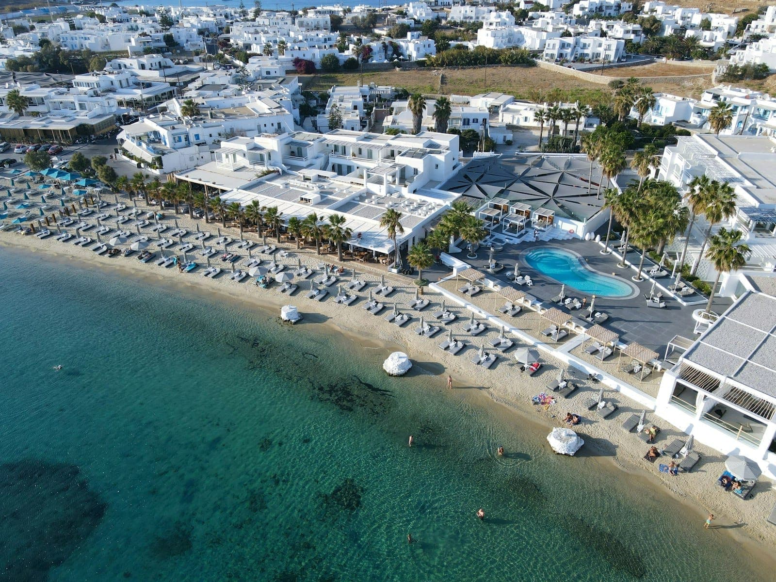 Luftaufnahme eines Beach Clubs auf Mykonos mit Liegestühlen, Sonnenschirmen, einem Pool und weiß getünchten Gebäuden direkt am türkisfarbenen Wasser.