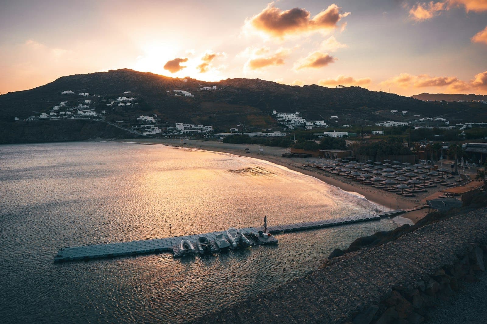 Sonnenuntergang über einem Beach Club auf Mykonos mit Reihen von Liegestühlen, ruhigem Wasser und an einem Pier festgemachten Booten, der die Sommersaison der Insel heraufbeschwört.