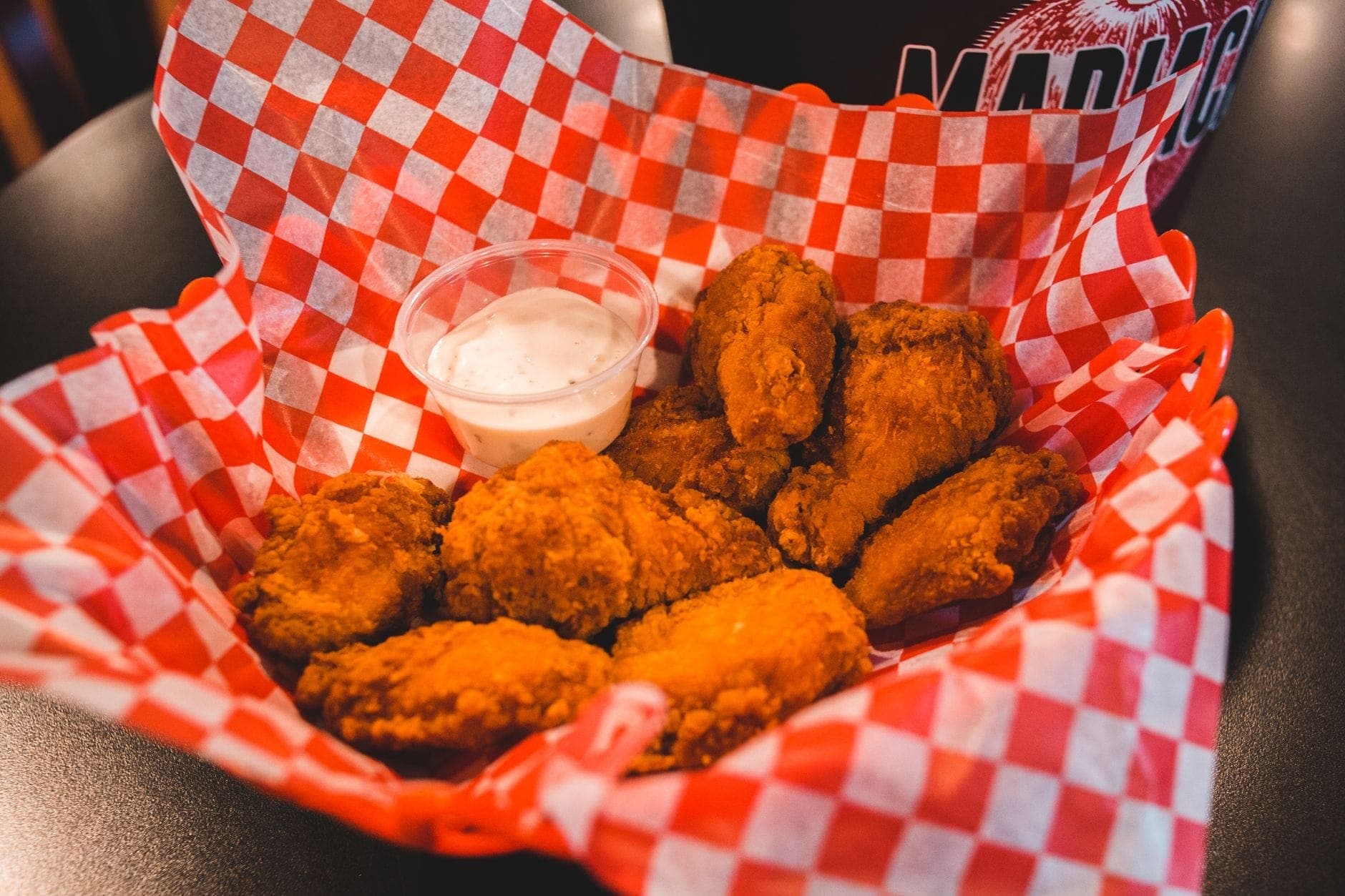 Ein Korb mit Nashville-Style Hot Chicken Wings mit Dip-Sauce auf einer rot karierten Unterlage an einem Restauranttisch.