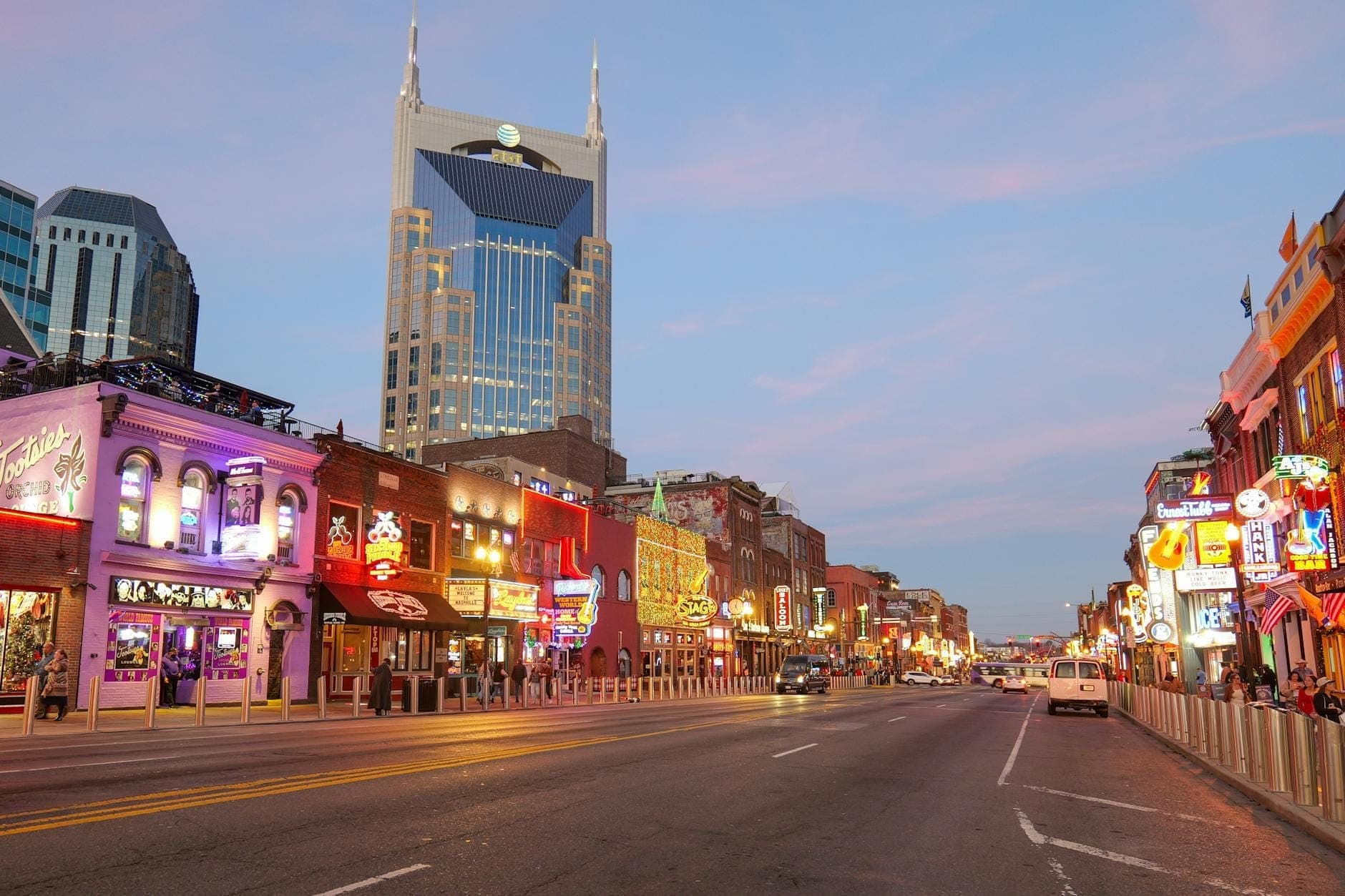 Broadway w centrum Nashville o zachodzie słońca, z kolorowymi barami, neonami i kultowym budynkiem AT&T w tle.