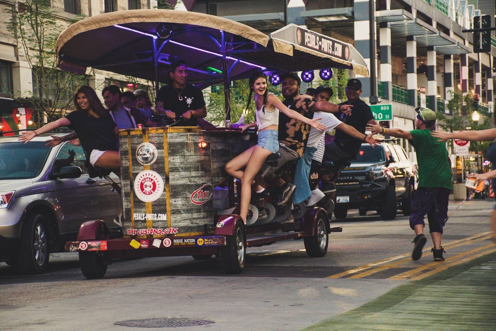 Ożywiona grupa ludzi bawiąca się podczas przejażdżki pedal tavern z neonowymi światłami, poruszająca się przez ruchliwą ulicę w miejskim otoczeniu.