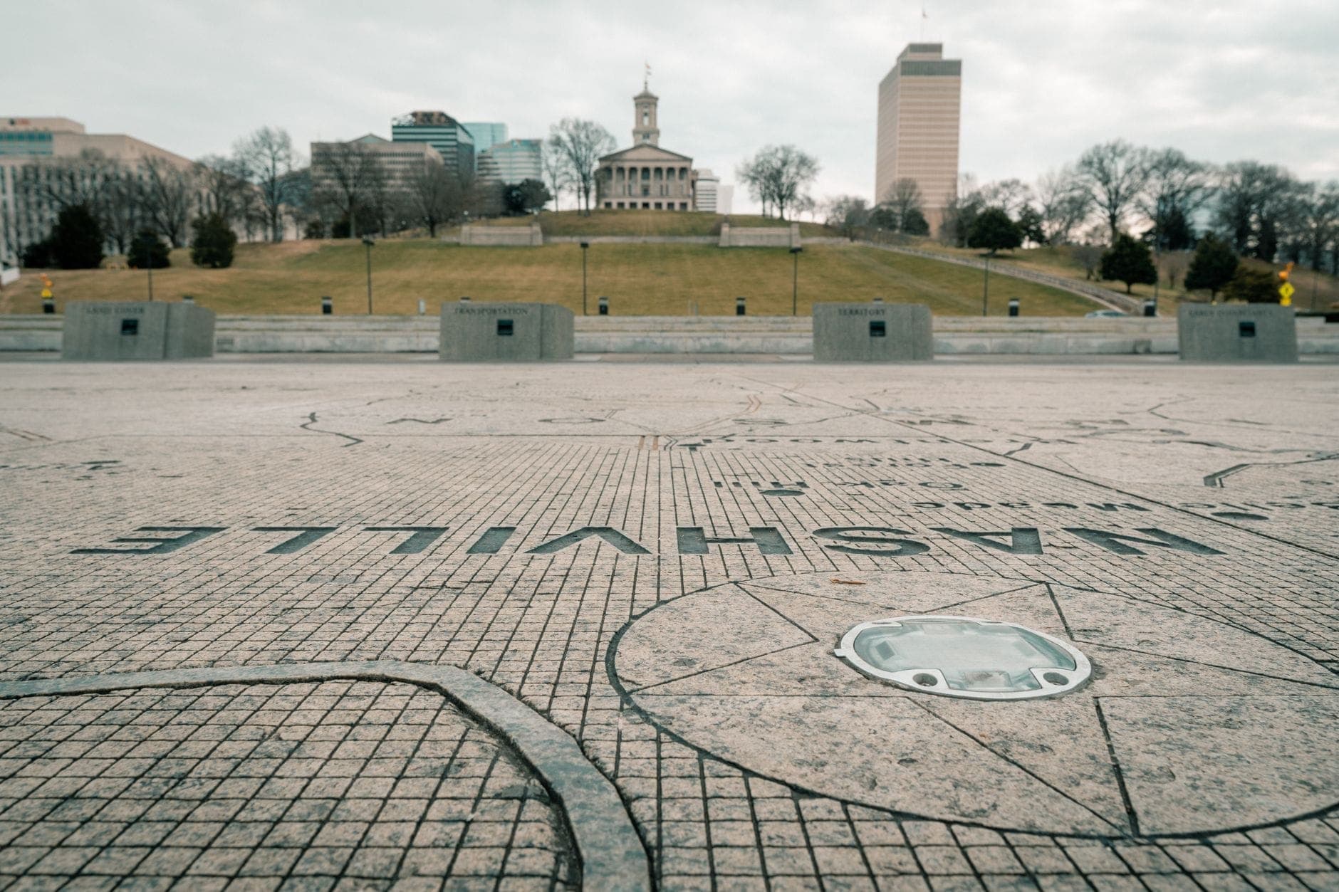View of the Tennessee State Capitol on a hill, seen from Bicentennial Park with a stone map of Nashville in the foreground under a cloudy sky.