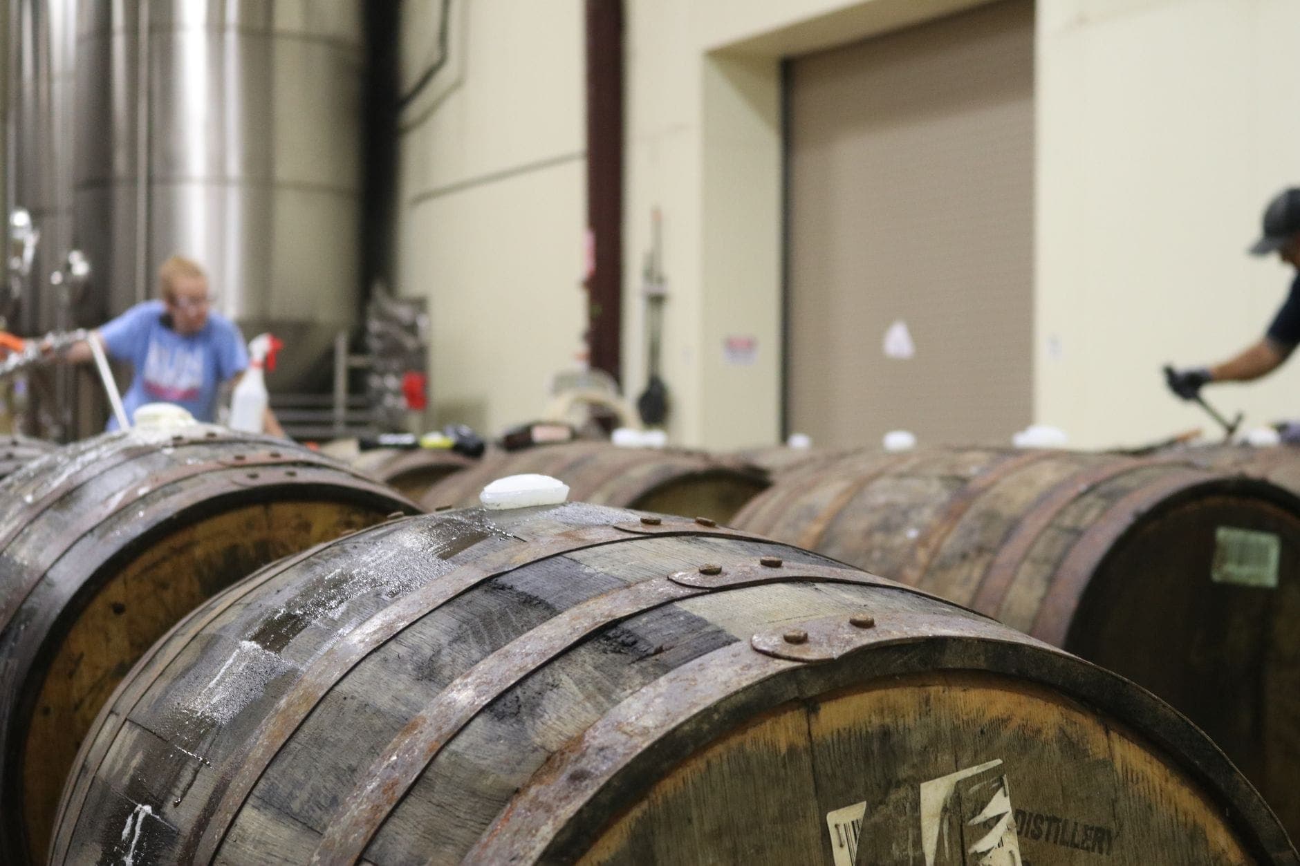 Rzędy beczek whiskey wewnątrz magazynu destylarni, z pracownikami w tle zajmującymi się beczkami.