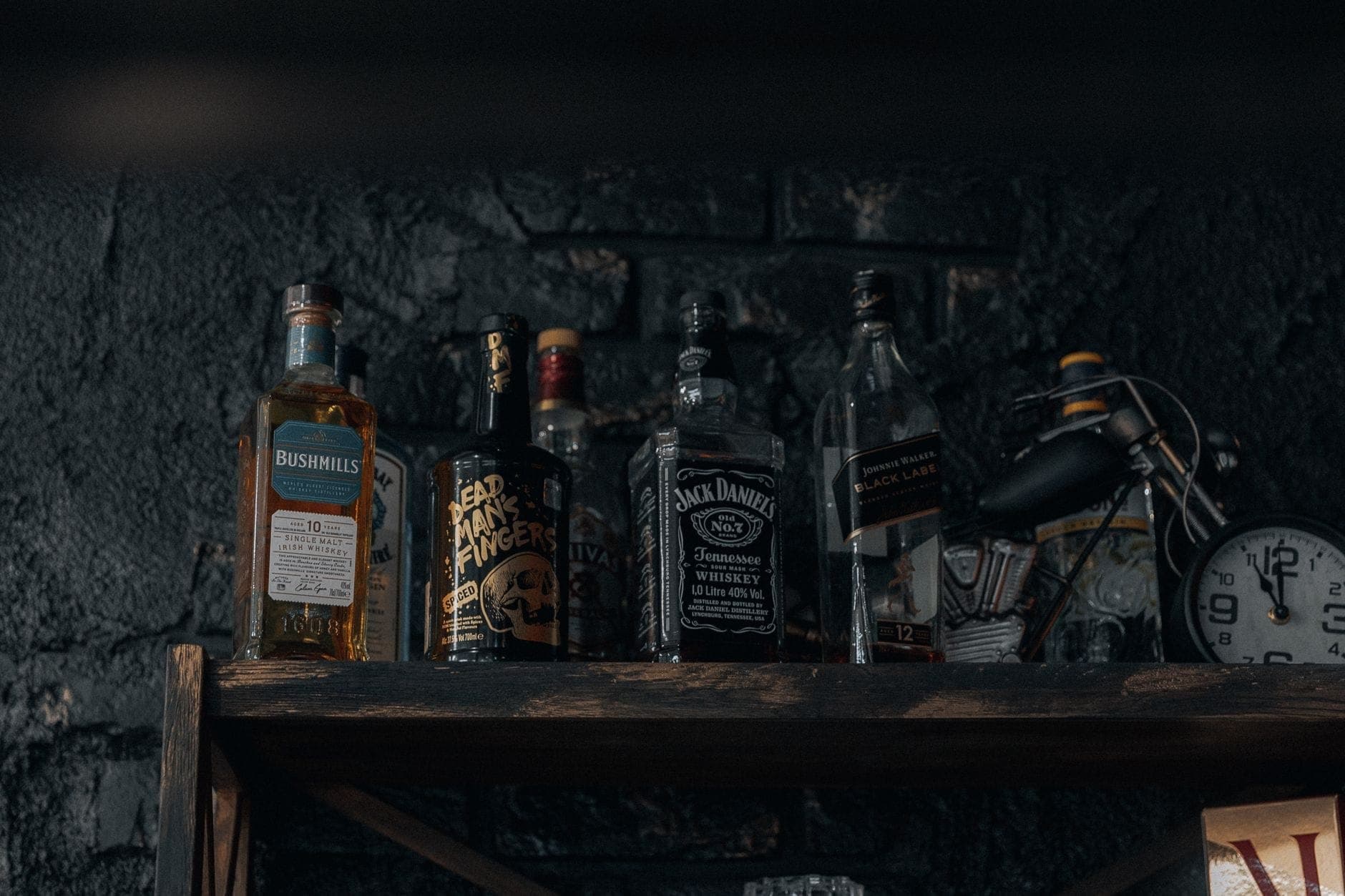 Półka z butelkami whisky, w tym Jack Daniel's, na ciemnym, rustykalnym tle, z zegarem i małymi dekoracyjnymi przedmiotami w pobliżu.