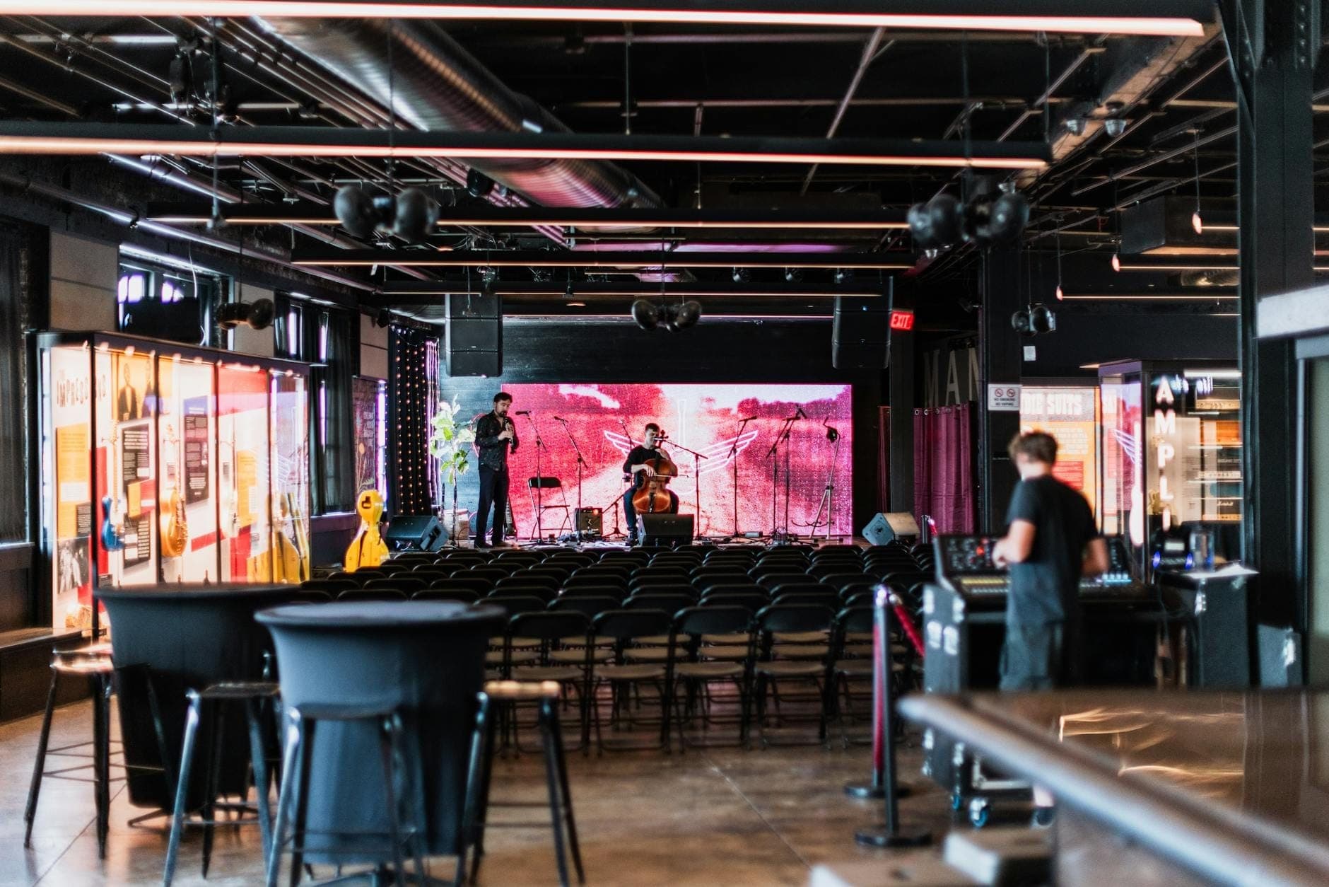 Local moderno de música ao vivo com um pequeno palco, cadeiras vazias e alguns músicos se preparando, provavelmente em um bairro de Nashville.