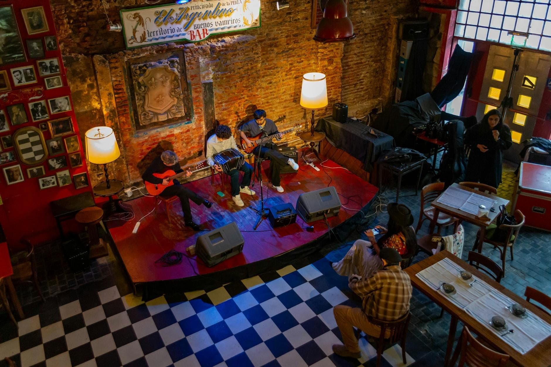 Pequeno grupo de músicos se apresentando em um palco aconchegante em um local com paredes de tijolo enquanto o público sentado ouve atentamente.