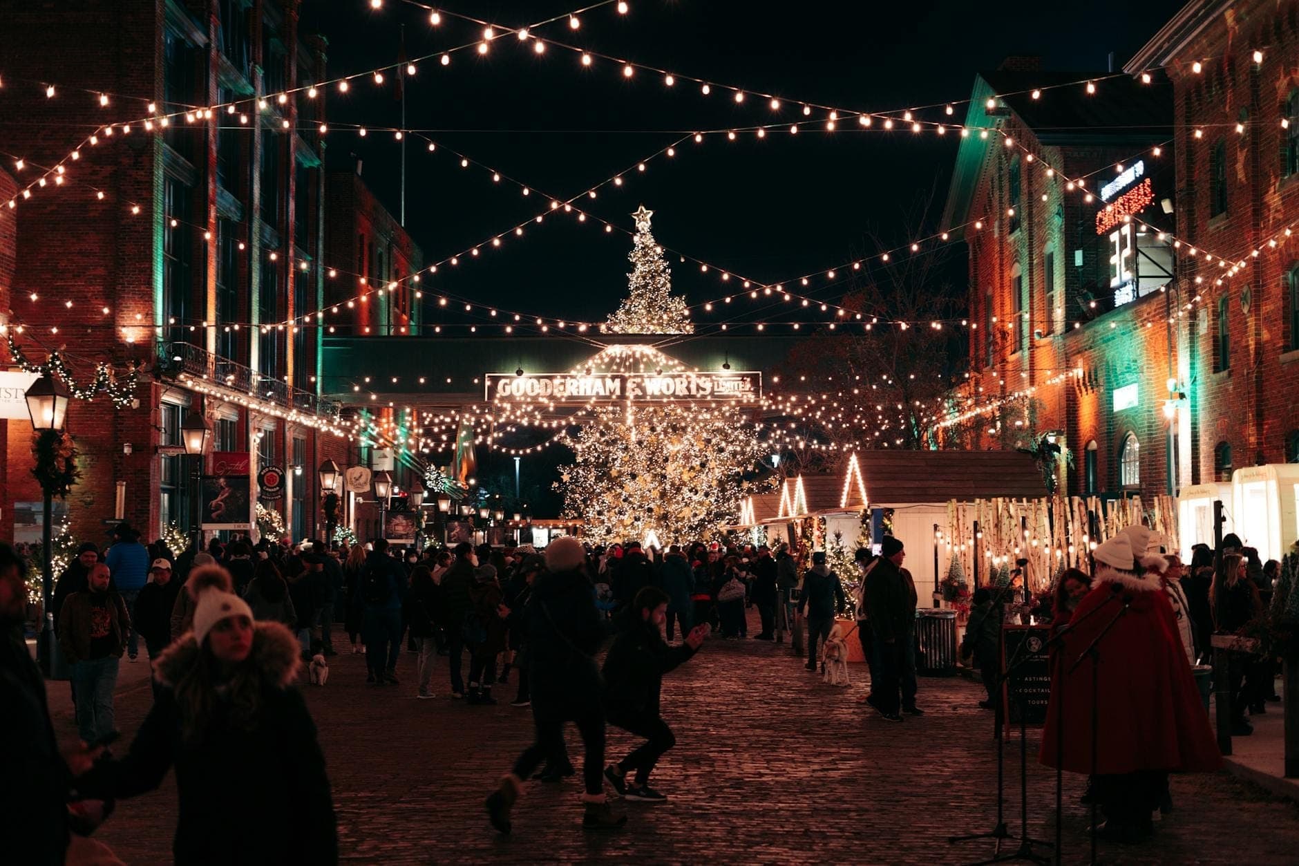 Festive outdoor market scene at night with people walking under string lights, a large illuminated Christmas tree, and historic brick buildings decorated for the holidays.