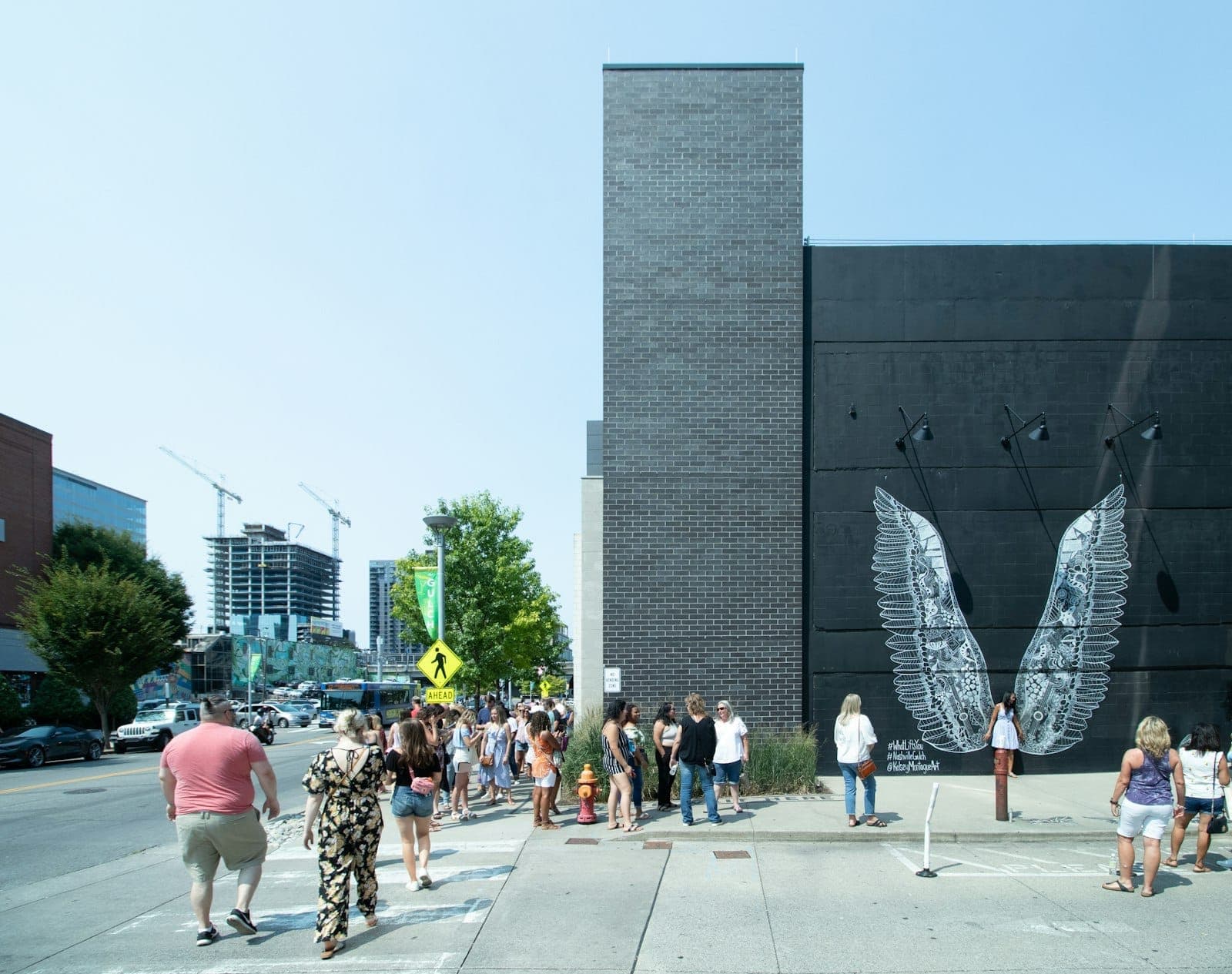 Wide street view of the iconic black mural with white wings in Nashville, with people waiting in line to take photos and city buildings in the background.
