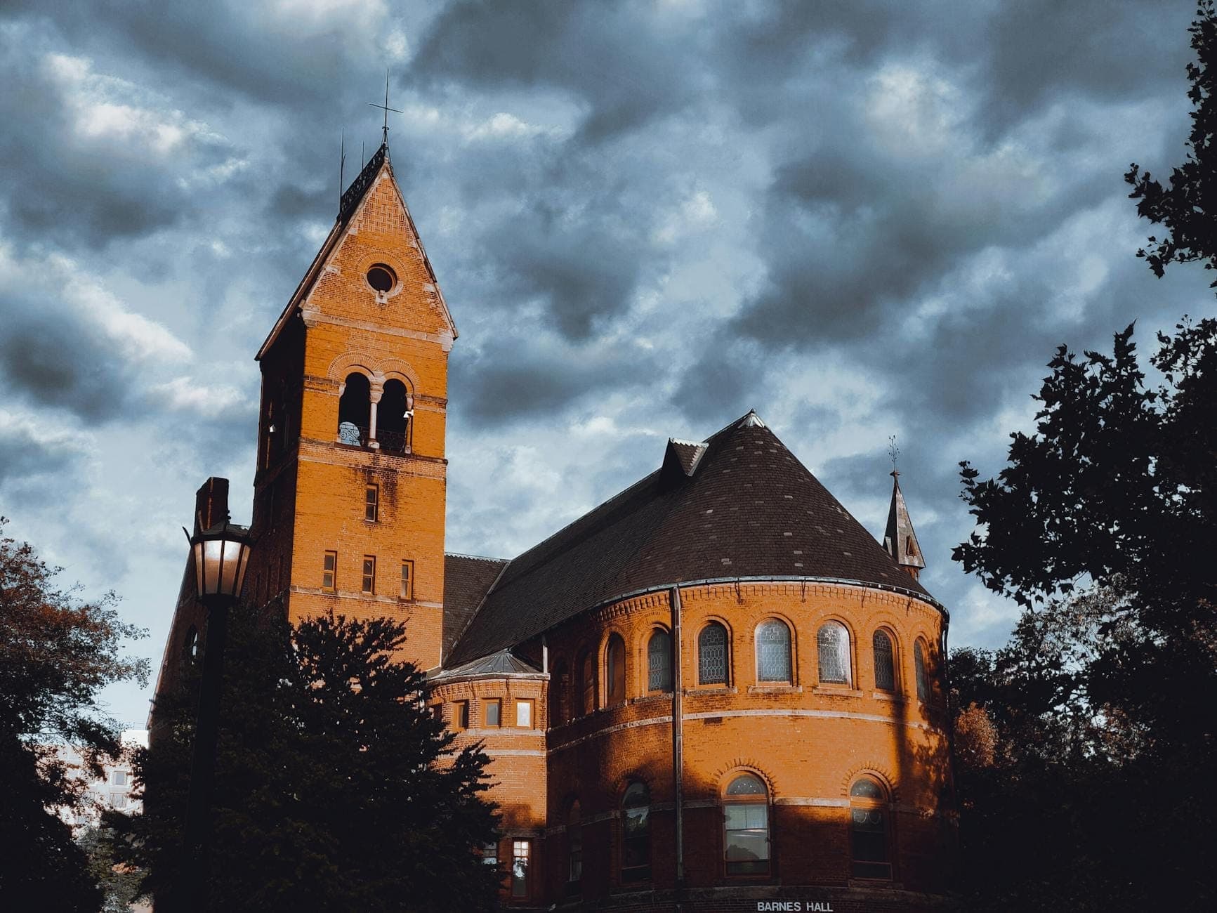 Histórico edificio universitario de ladrillo rojo con una alta torre campanario y ventanas en arco bajo un cielo dramático, rodeado de árboles.