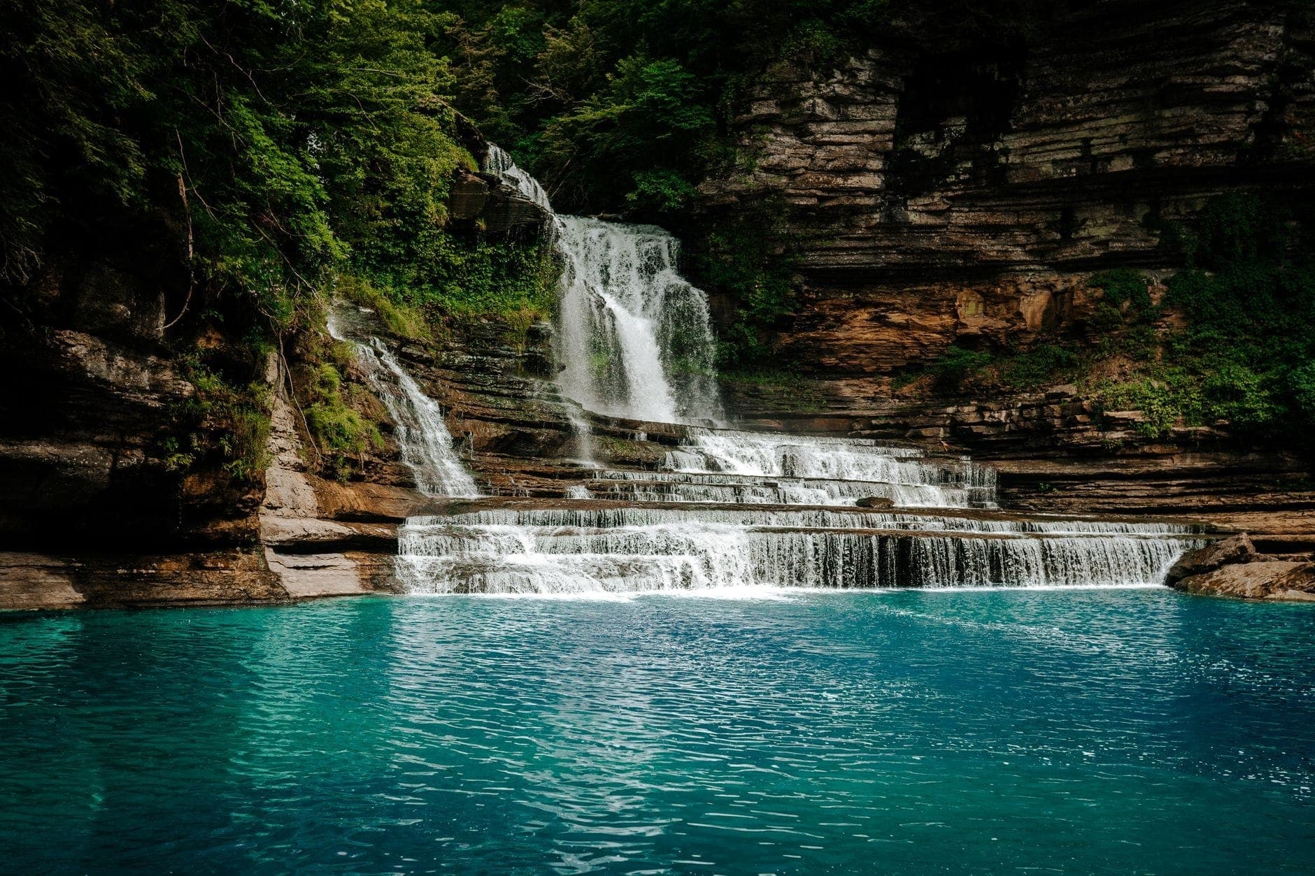 Wide waterfall cascading over rocky ledges into a clear blue pool, surrounded by lush green forest in a scenic state park setting.