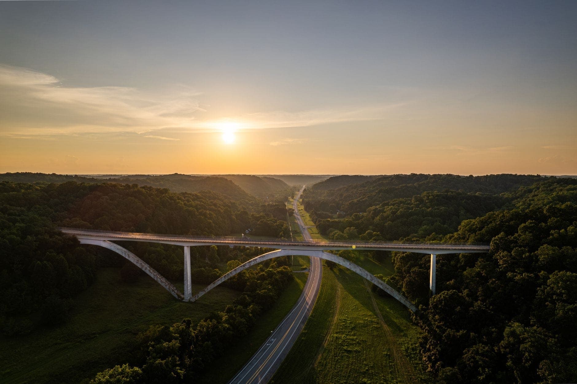 A sweeping aerial view of a scenic highway bridge surrounded by lush green hills at sunset, evoking the start of a beautiful Tennessee road trip.