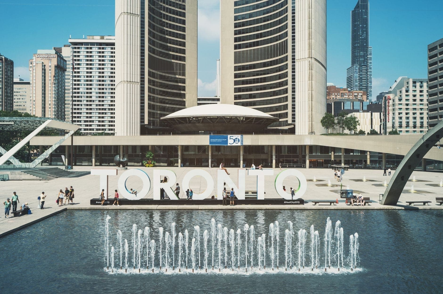 Vista diurna di Nathan Phillips Square con il Municipio di Toronto, l'insegna TORONTO, la fontana e persone che si godono la piazza davanti all'edificio modernista.