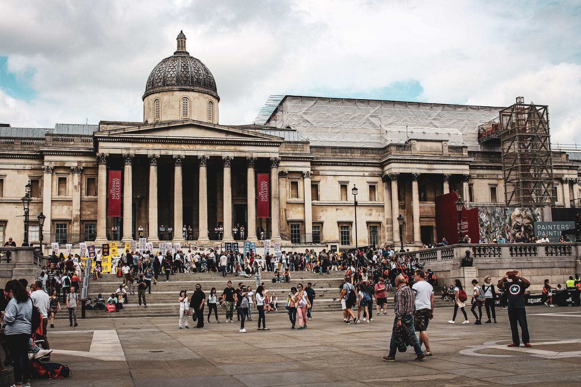 Szeroki widok na National Gallery w Londynie z okazałą fasadą, centralną kopułą i tłumem odwiedzających przed wejściem na Trafalgar Square pod zachmurzonym niebem.