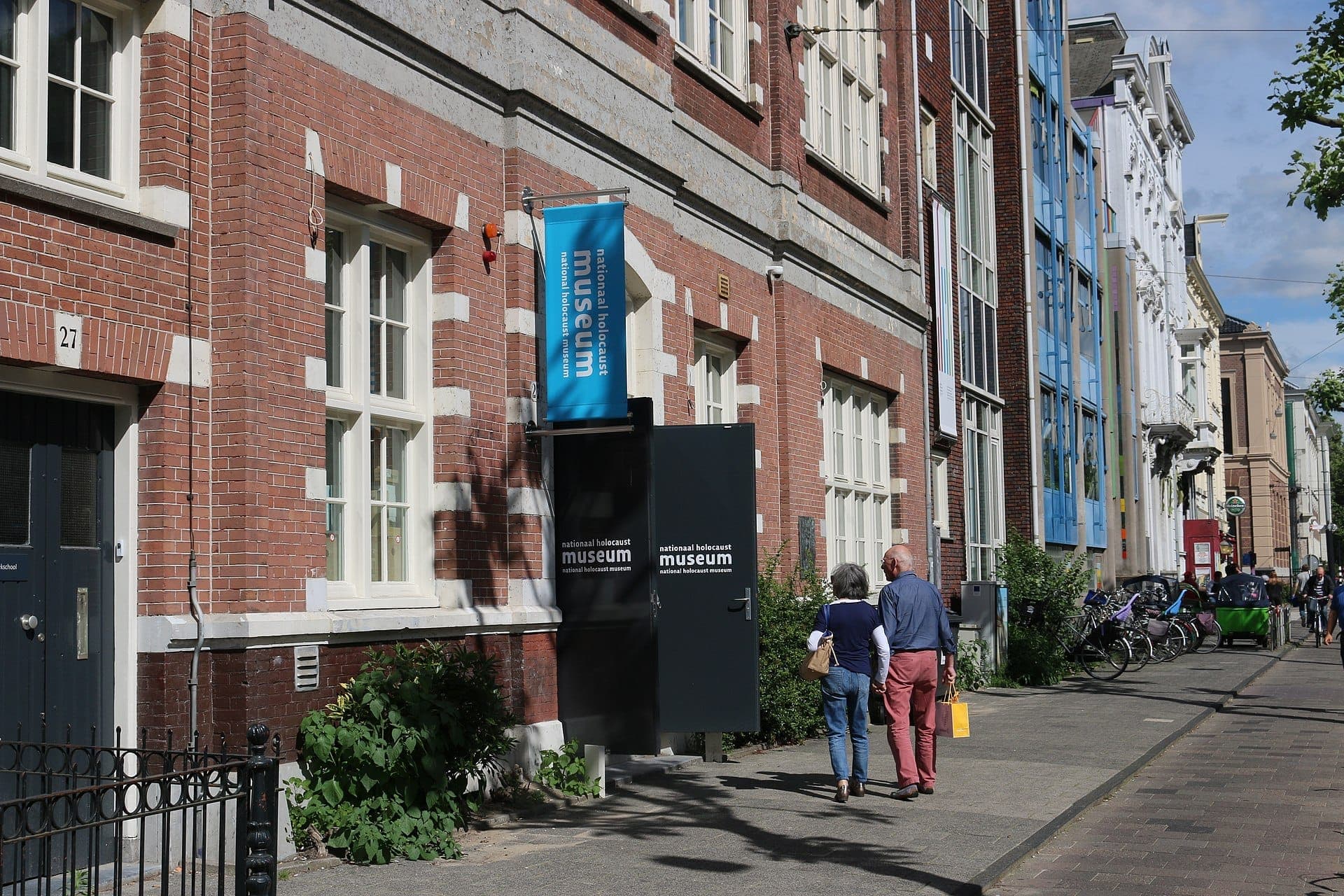 Außenansicht des National Holocaust Museum Amsterdam mit seiner roten Backsteinsfassade, blauer Beschilderung und zwei vorbeigehenden Personen an einem sonnigen Tag.