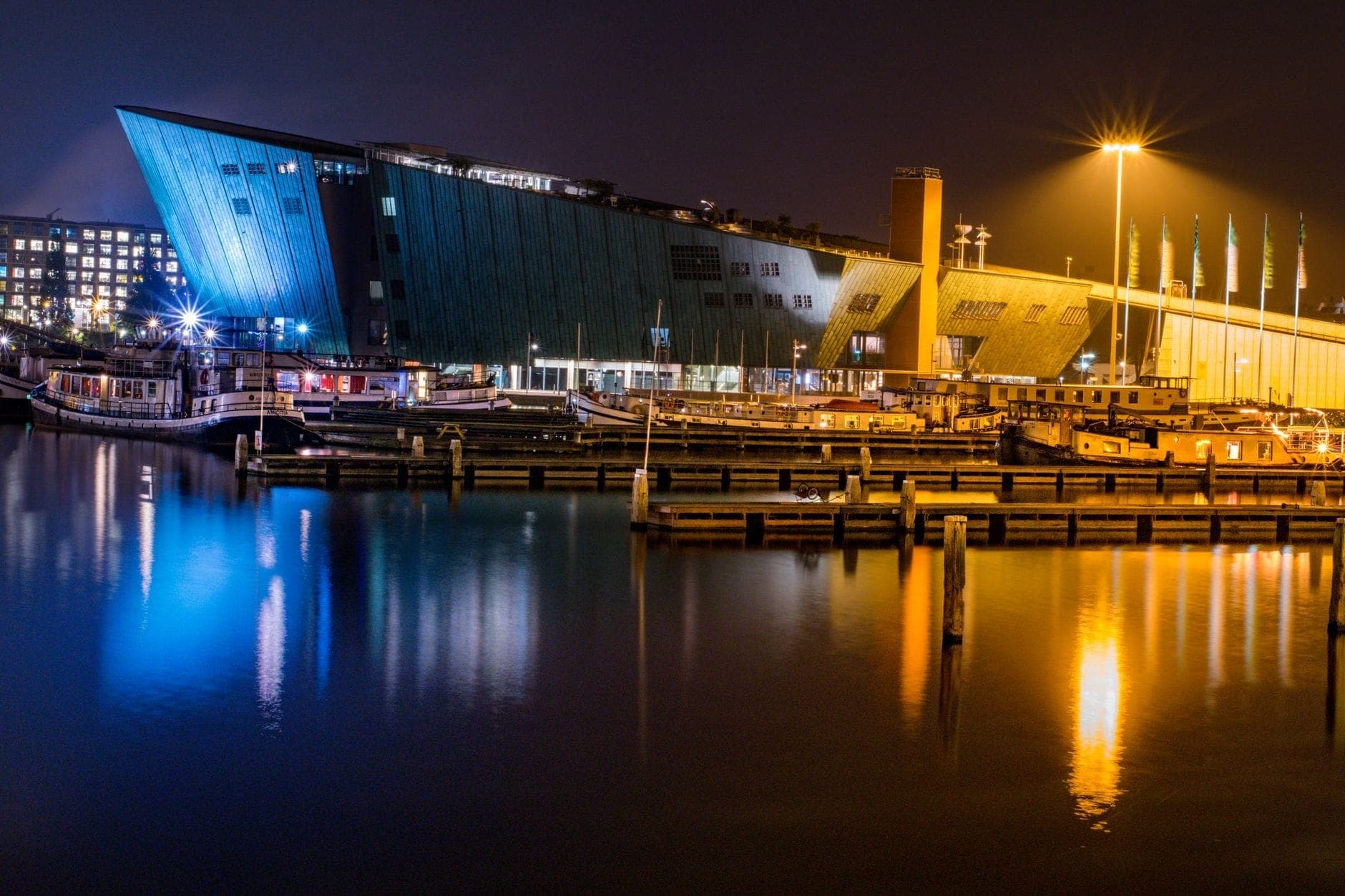 Nachtansicht eines modernen Schifffahrtsmuseums am Wasser mit markanter schiffsartiger Architektur, die sich im ruhigen Hafenbecken Amsterdams spiegelt.