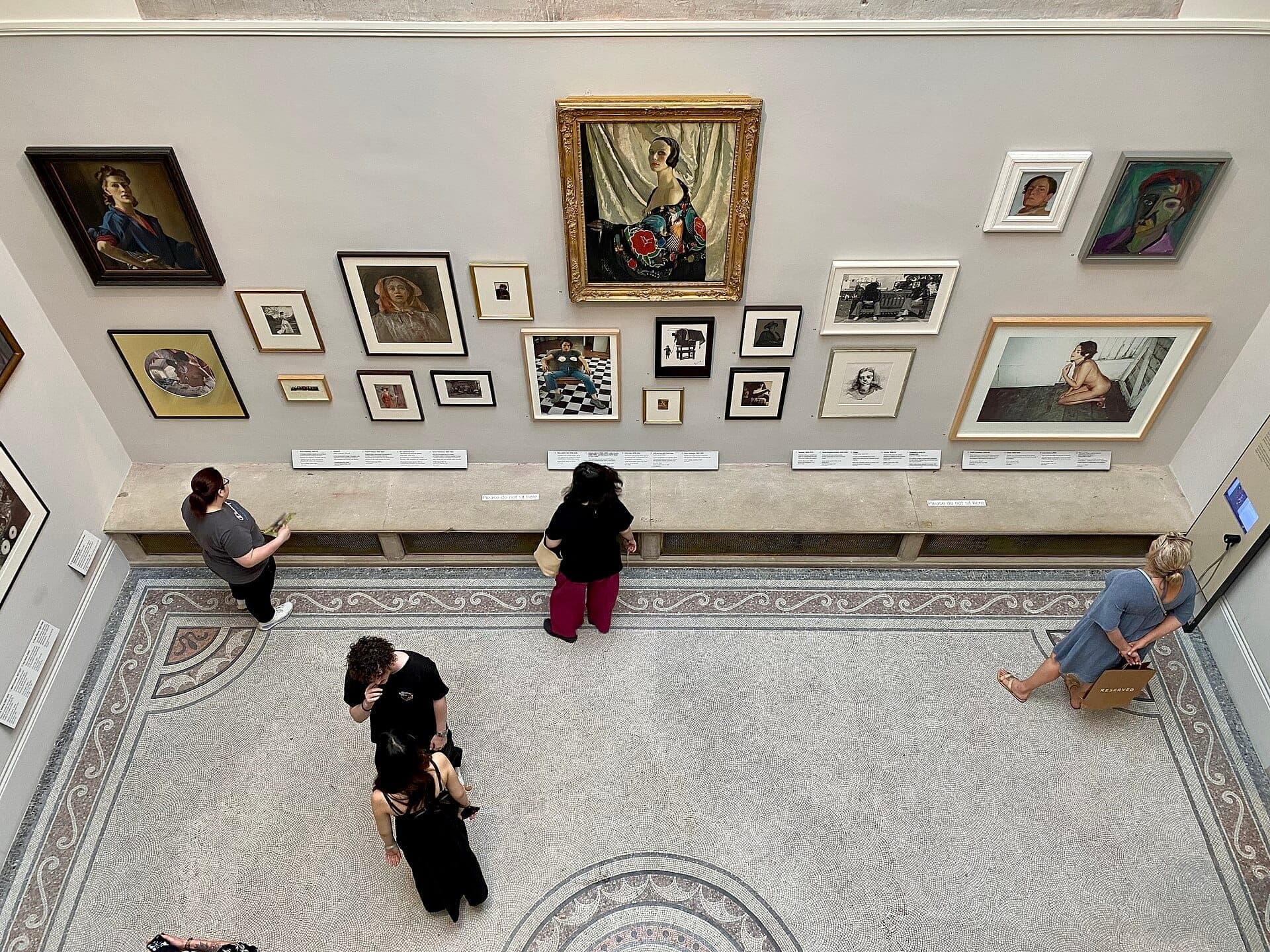 Besucher betrachten eine eklektische Sammlung gerahmter Porträts, die an einer hell gefärbten Wand in der National Portrait Gallery London ausgestellt sind.
