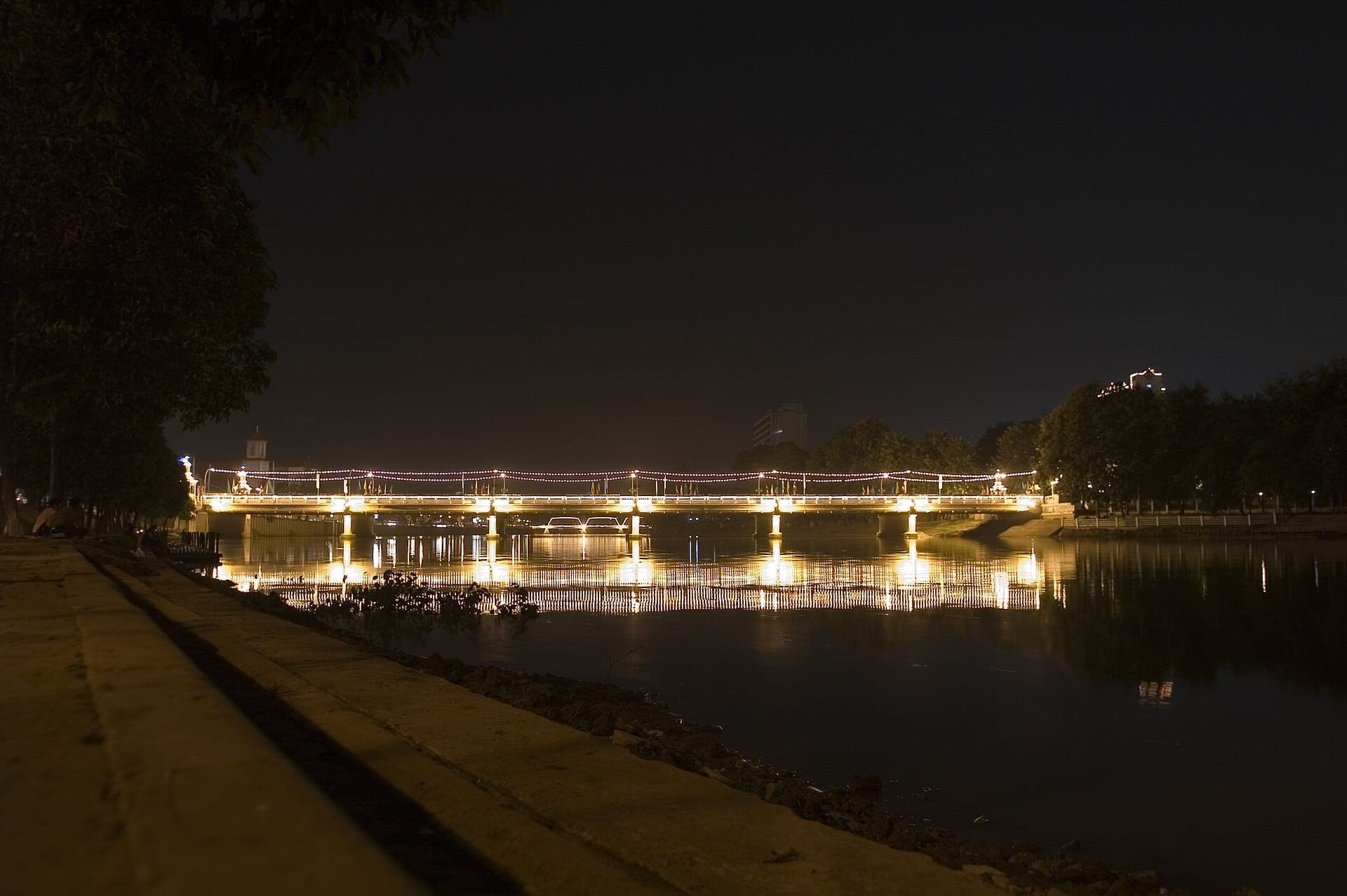Die Nawarat-Brücke bei Nacht beleuchtet, mit Lichtern, die sich im ruhigen Ping-Fluss spiegeln, baumgesäumten Ufern und Gebäuden in der Ferne.