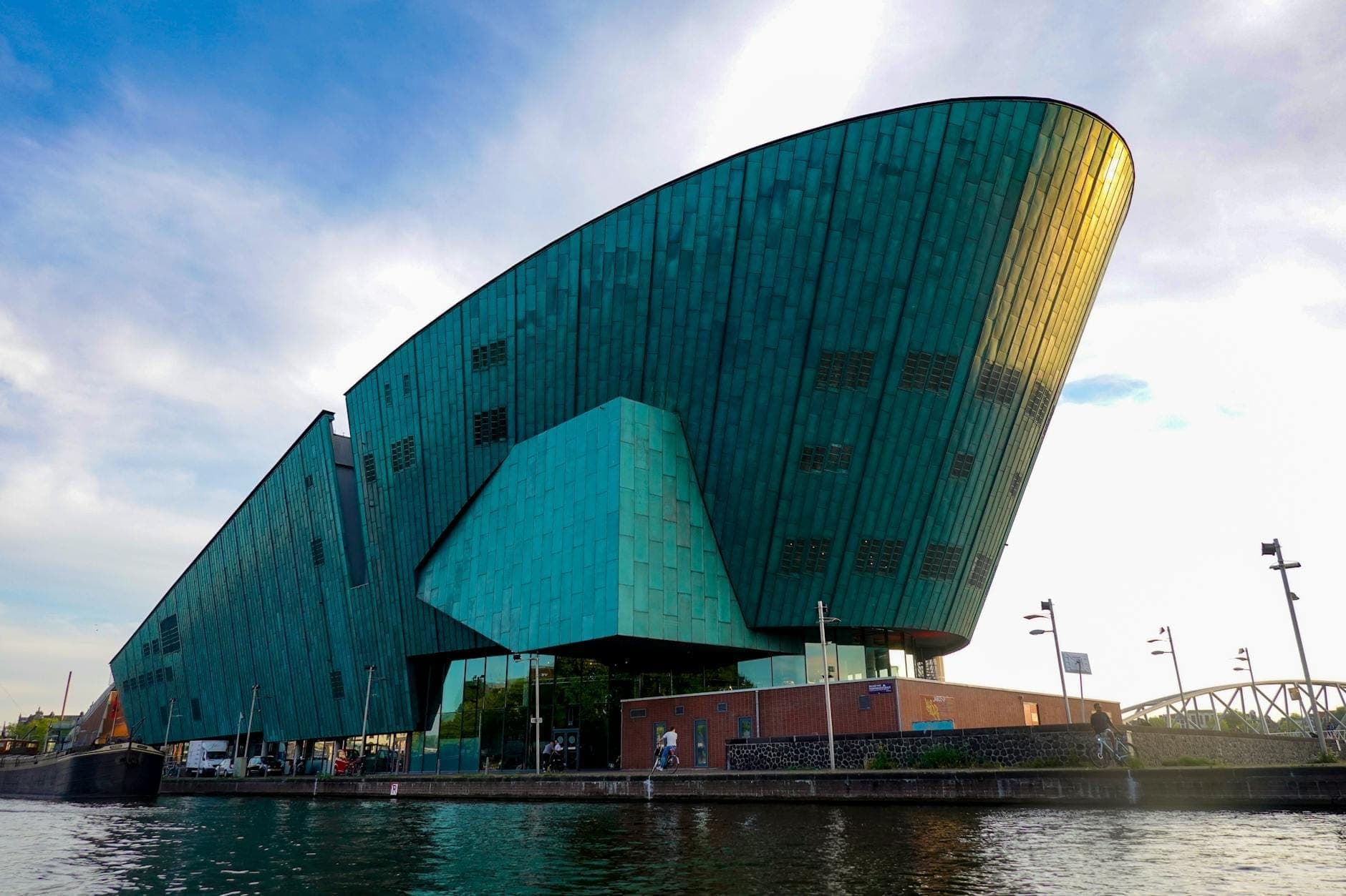 Das markante, grün-kupferfarbene NEMO Science Museum vom Hafenufer aus gesehen, mit blauem Himmel und Menschen beim Eingang.
