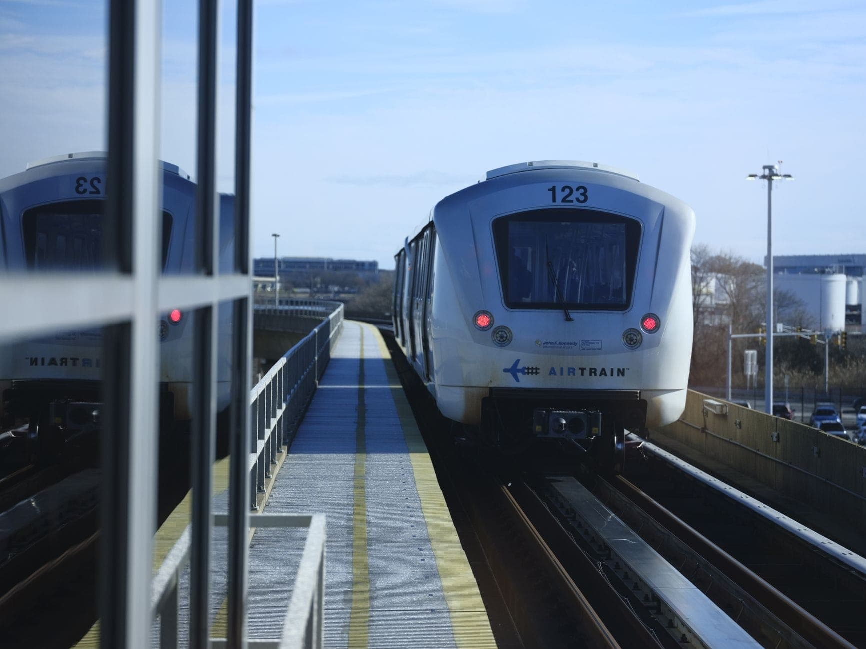 JFK AirTrain กำลังเข้าสู่ชานชาลาสถานียกระดับ มองเห็นรางรถไฟ แสงกลางวันสว่างจ้า บรรยากาศระบบขนส่งสมัยใหม่