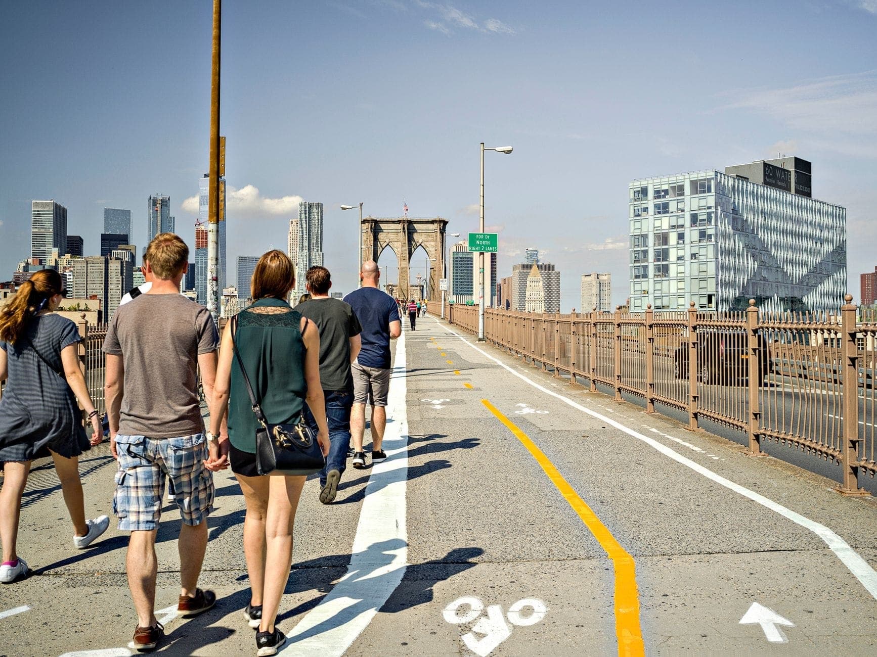 ผู้คนเดินบนทางเดินสำหรับคนเดินเท้าของ Brooklyn Bridge โดยมีตึกระฟ้าของ Manhattan เป็นฉากหลังในวันที่แดดจัด