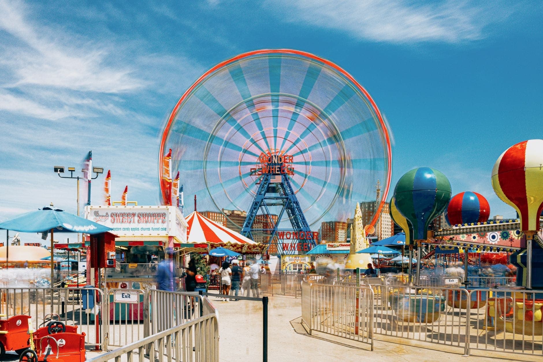 วิวสดใสของสวนสนุก Coney Island แสดงให้เห็น Wonder Wheel เครื่องเล่นในงานคาร์นิวัล บูธสีสันสดใส และผู้คนที่เพลิดเพลินกับบอร์ดวอล์กใต้ท้องฟ้าสีฟ้า