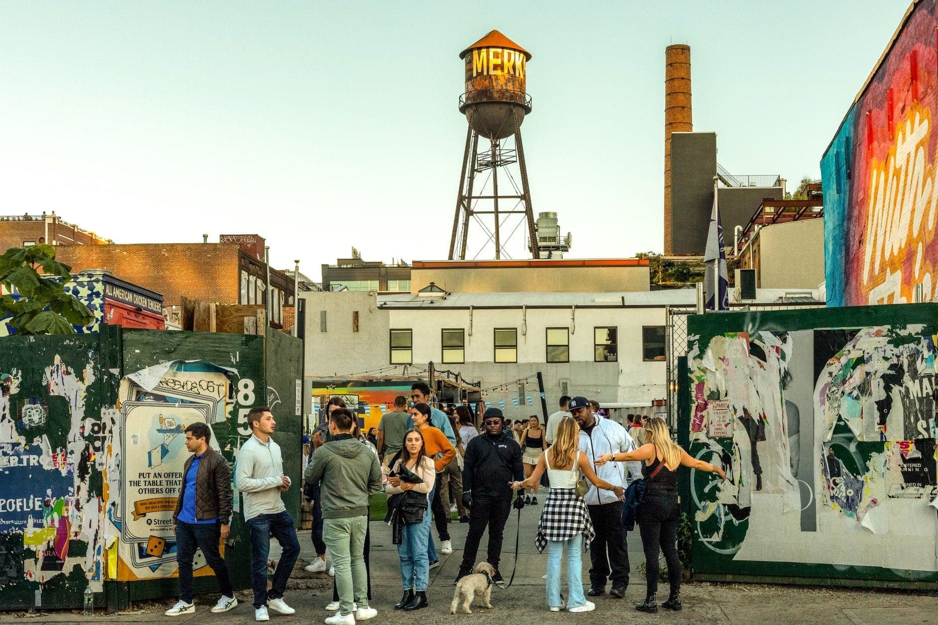 กลุ่มคนหนุ่มสาวที่มารวมตัวกันใกล้แผงตลาดสีสันสดใสใน Williamsburg โดยมีหอเก็บน้ำอันเป็นสัญลักษณ์และงานศิลปะริมถนนอยู่เบื้องหลัง