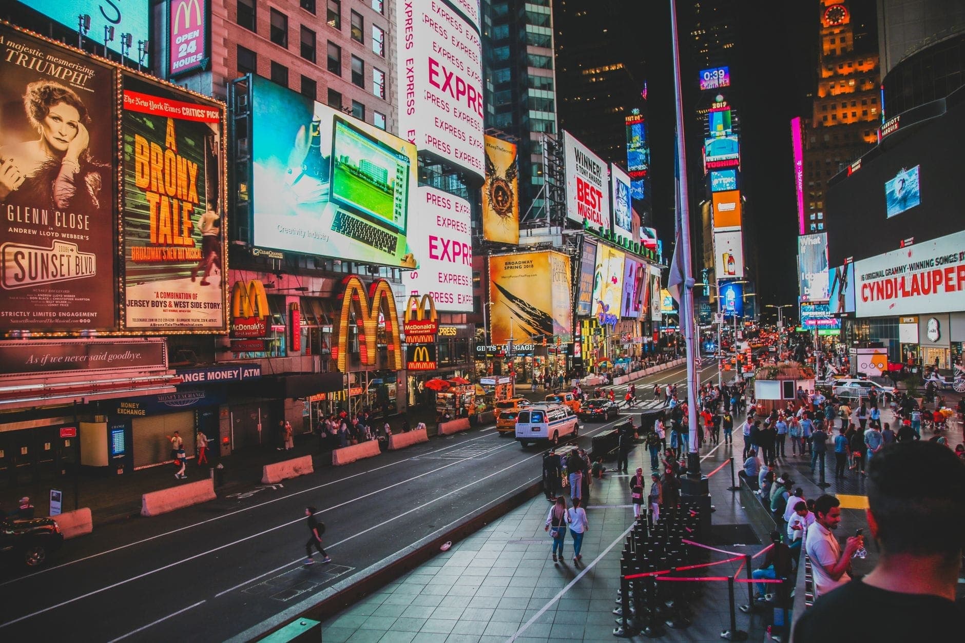 Times Square ที่คึกคักในยามค่ำคืน พร้อมป้ายโฆษณาสีสันสดใส ฝูงชนบนท้องถนน และแสงไฟจ้า สะท้อนบรรยากาศอันมีชีวิตชีวาของ Midtown ใน New York City
