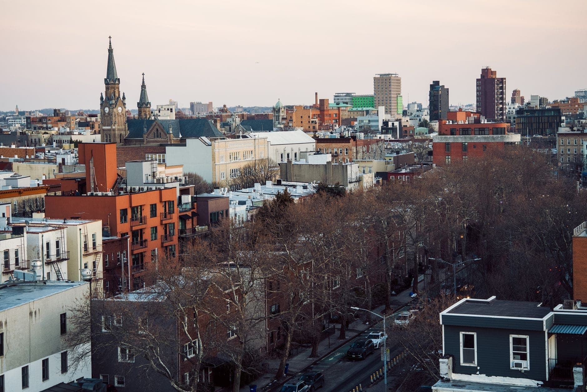 มุมมองทางอากาศของเขตหนึ่งใน New York City ที่มีอาคารเตี้ย โบสถ์ และถนนที่เรียงรายไปด้วยต้นไม้ในยามพลบค่ำ
