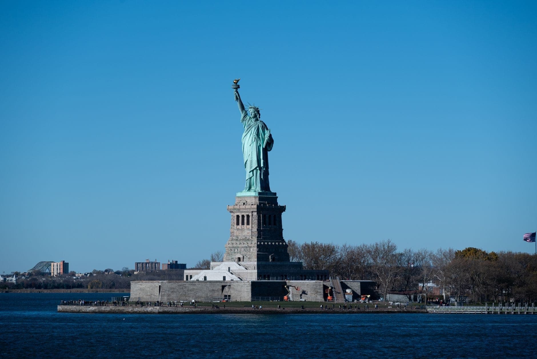 วิวกว้างของ Statue of Liberty บนเกาะในเวลากลางวันที่สว่างจ้า ล้อมรอบด้วยน้ำ พร้อมท้องฟ้าสีฟ้าใสเป็นฉากหลัง