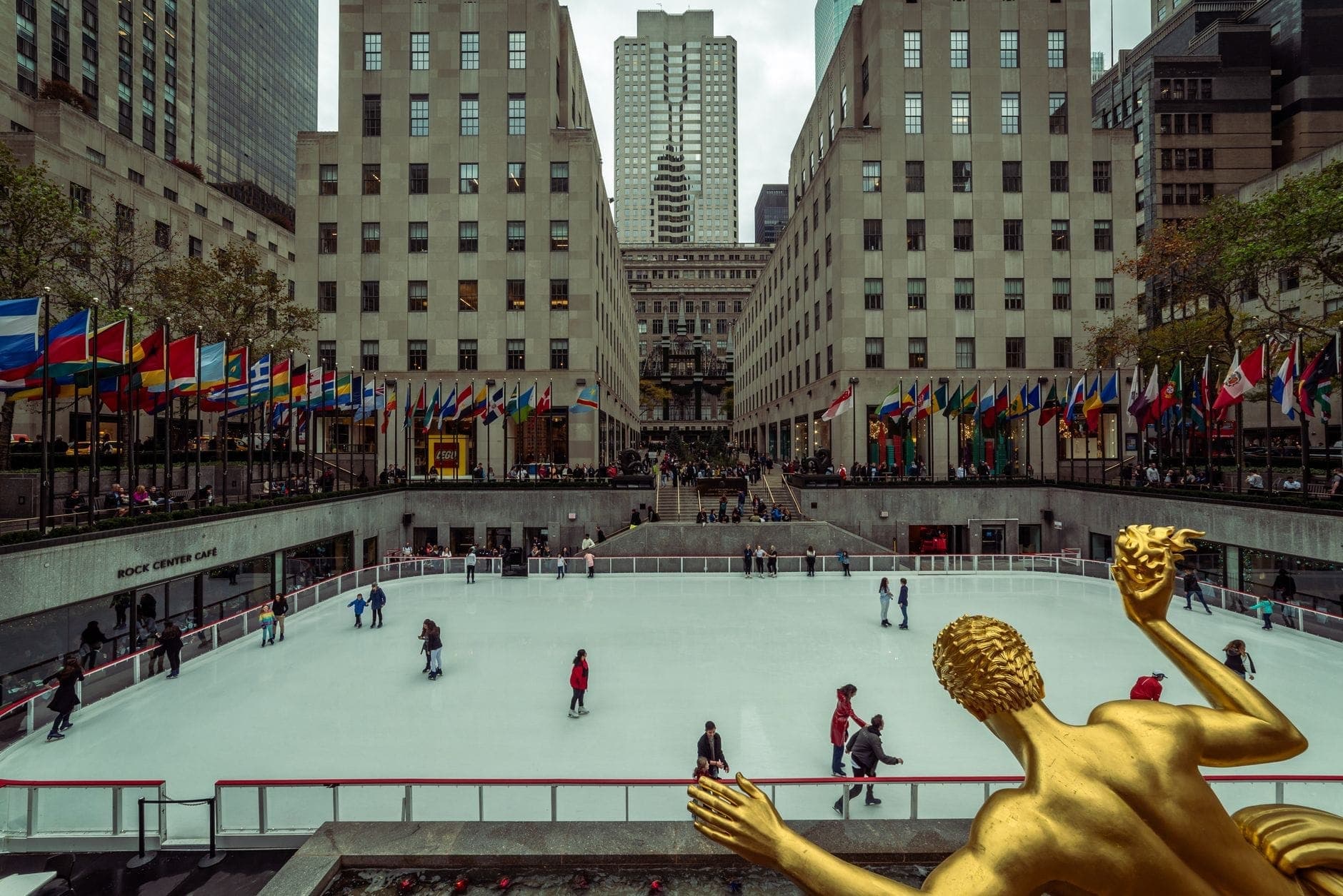 วิวกลางวันของลานสเก็ตน้ำแข็ง Rockefeller Center พร้อมนักสเก็ต รูปปั้น Prometheus สีทอง ธงชาติ และอาคารโดยรอบในนิวยอร์กซิตี้