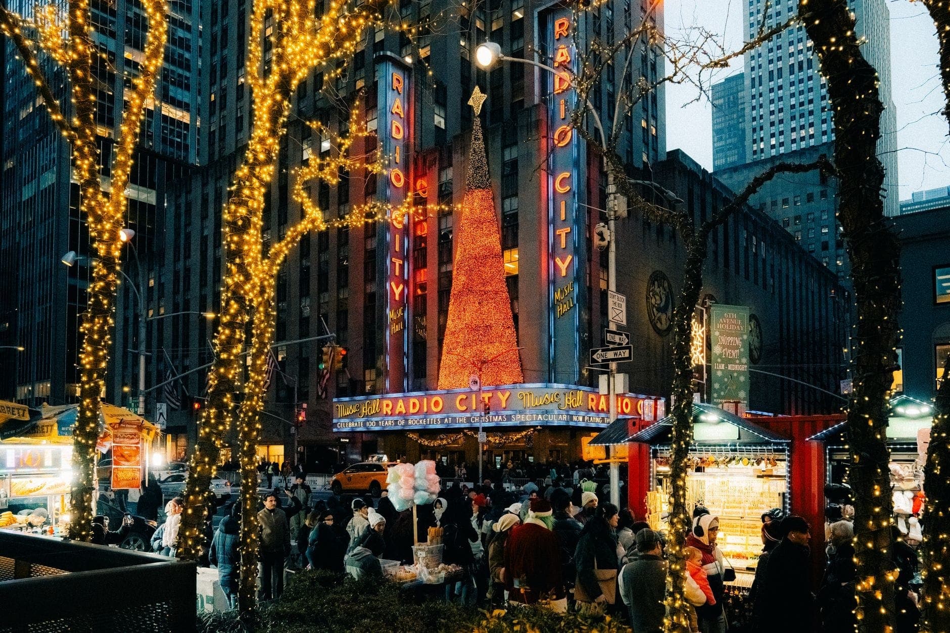Radio City Music Hall ในยามค่ำคืนช่วงเทศกาล พร้อมต้นคริสต์มาสขนาดใหญ่ที่สว่างไสวและไฟประดับเทศกาล มีฝูงชนมารวมตัวด้านนอก