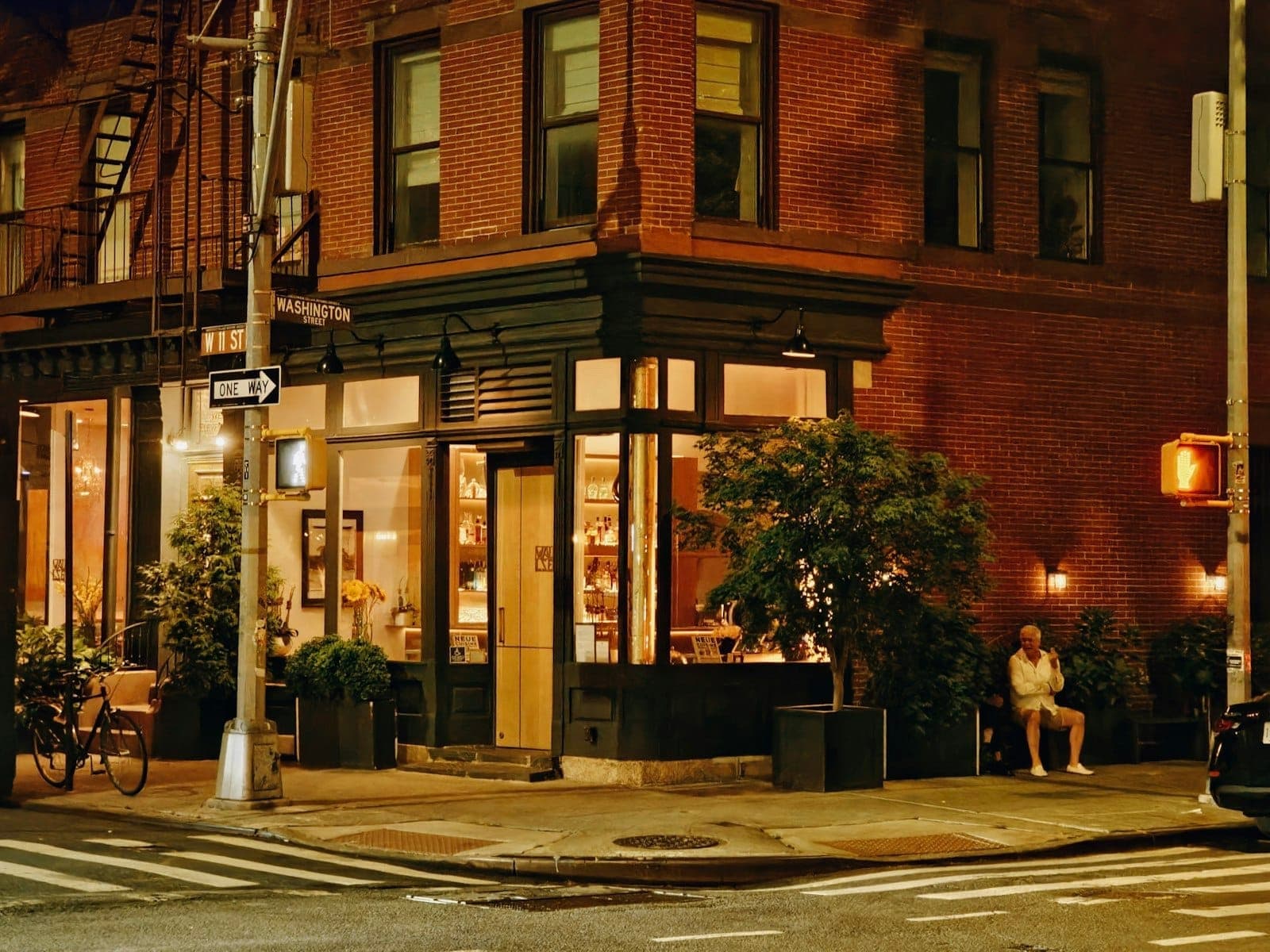 อาคารมุมที่มีแสงไฟอบอุ่นใน Greenwich Village ยามค่ำคืน มีคนนั่งอยู่ด้านนอก สถาปัตยกรรมอิฐคลาสสิกของ NYC