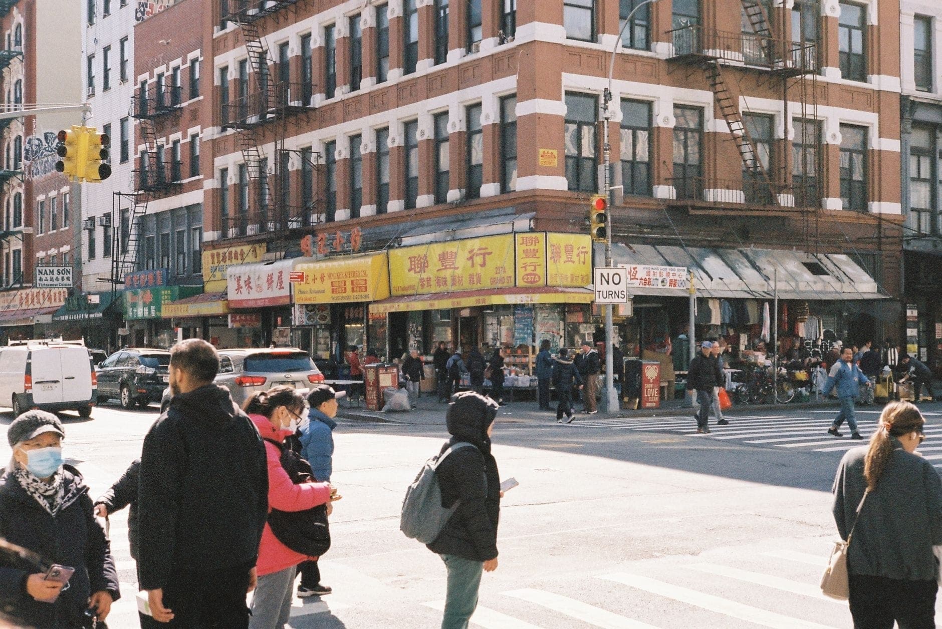 มุมถนนที่คึกคักใน New York City มีผู้คนข้ามถนน พร้อมด้วยร้านค้าและป้ายภาษาเอเชียในย่านเมืองที่หลากหลาย