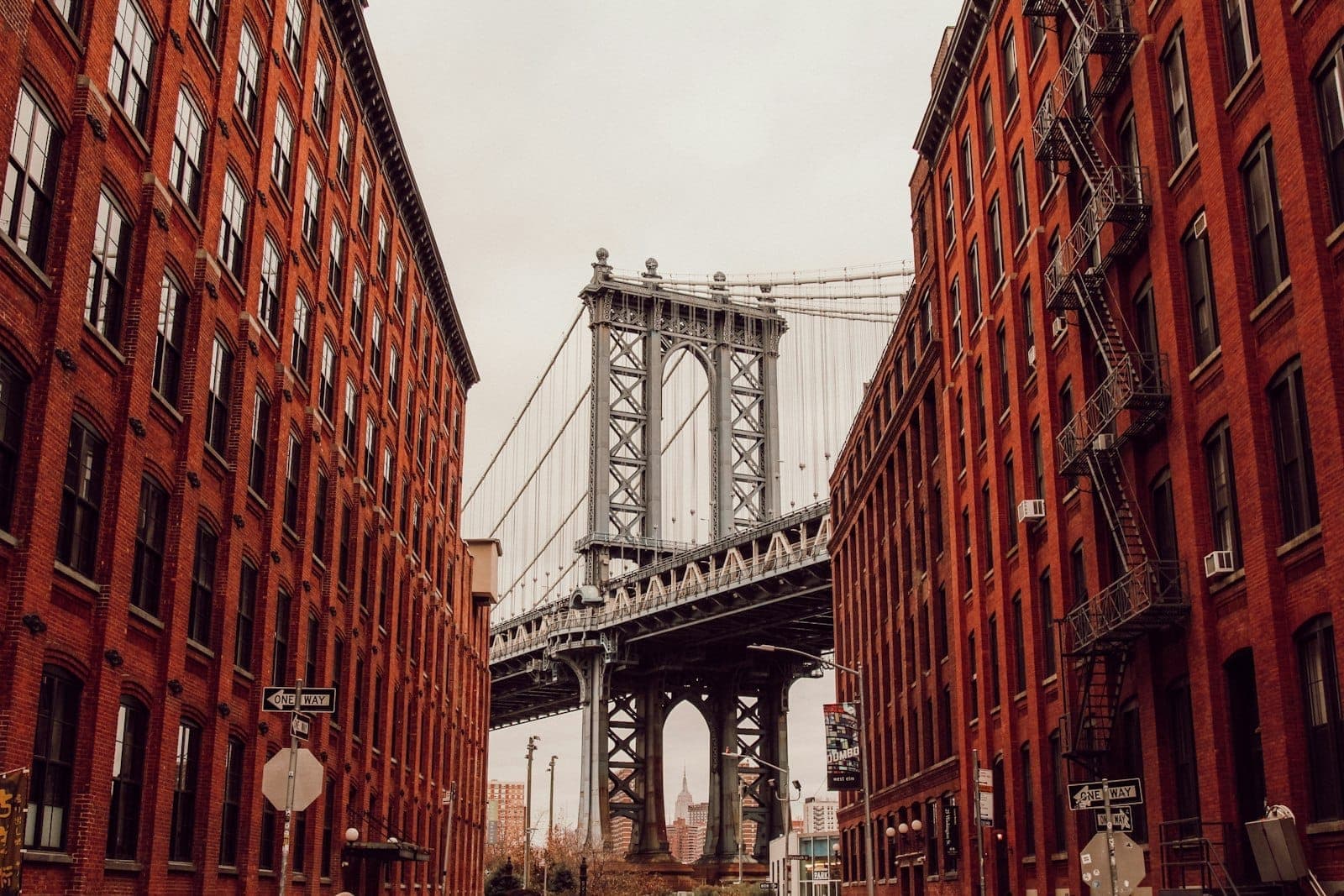 วิวของ Manhattan Bridge ที่ถูกโอบล้อมด้วยอาคารอิฐแดงใน DUMBO, Brooklyn ใต้ท้องฟ้าที่มีเมฆปกคลุม
