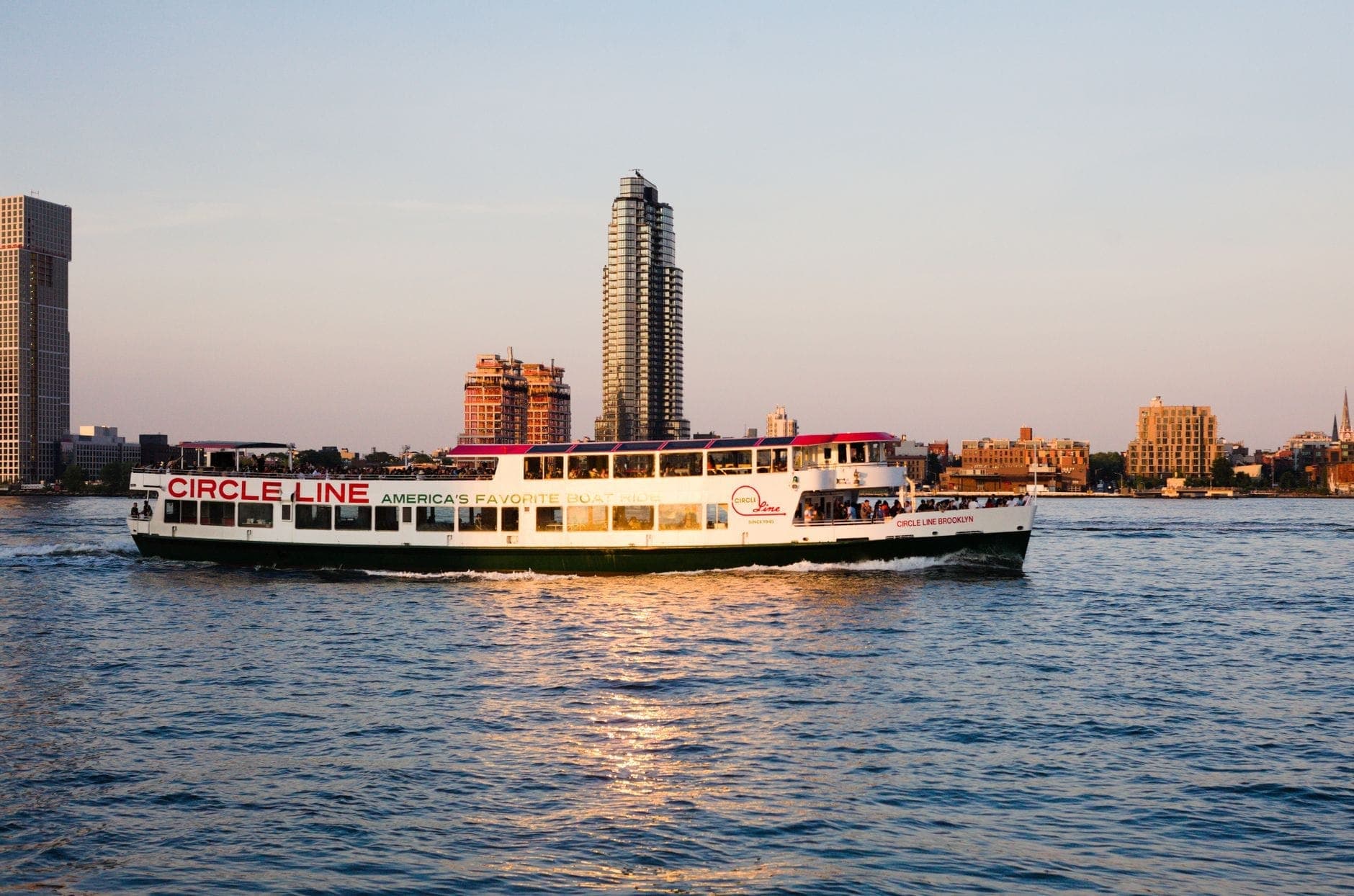 เรือล่องเรือปาร์ตี้ Circle Line บนน้ำพร้อมเส้นขอบฟ้าของ New York City เป็นฉากหลังยามพระอาทิตย์ตกดิน