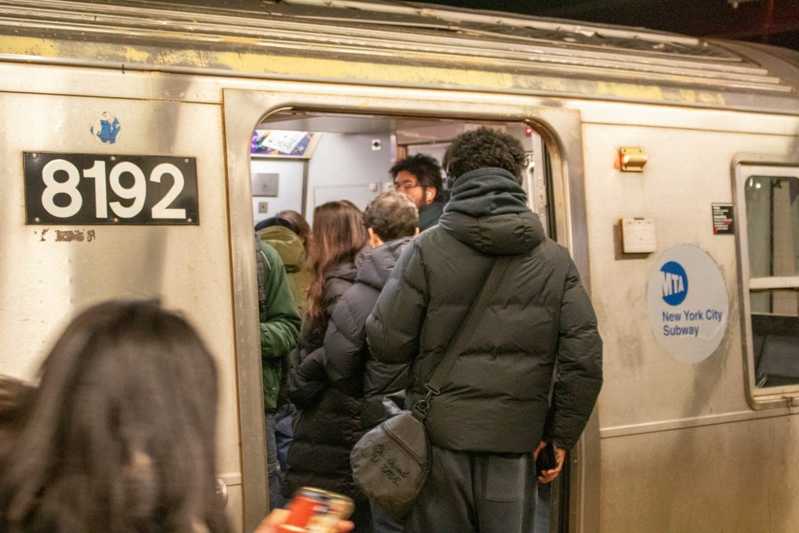 ฝูงชนกำลังขึ้นรถไฟใต้ดิน New York City ในยามค่ำคืน โดยมีโลโก้ MTA และหมายเลขรถไฟให้เห็นอย่างชัดเจน