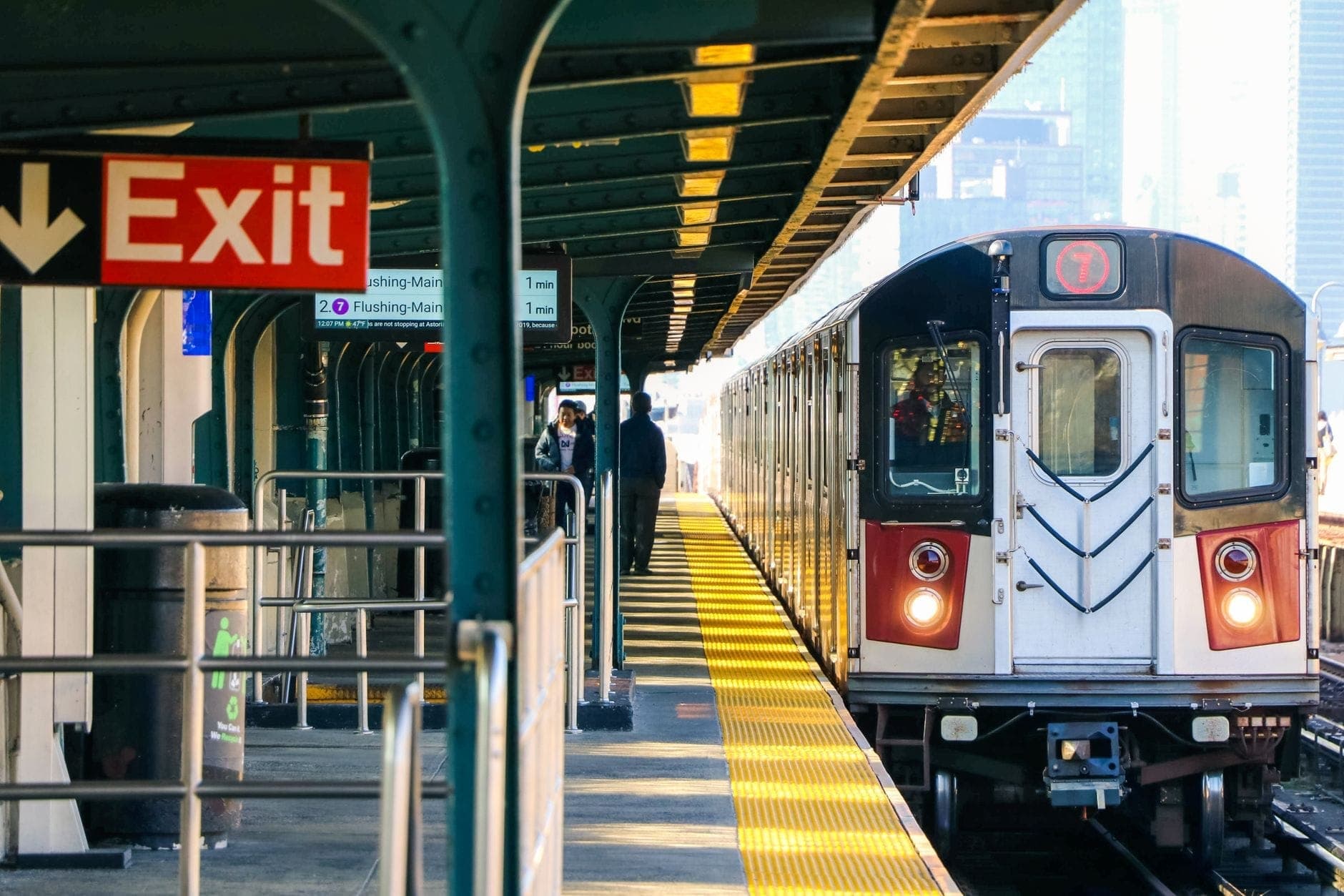 รถไฟสาย 7 ของ New York City กำลังเข้าสู่ชานชาลายกระดับพร้อมผู้โดยสารที่รอและป้ายทางออกที่มองเห็นได้