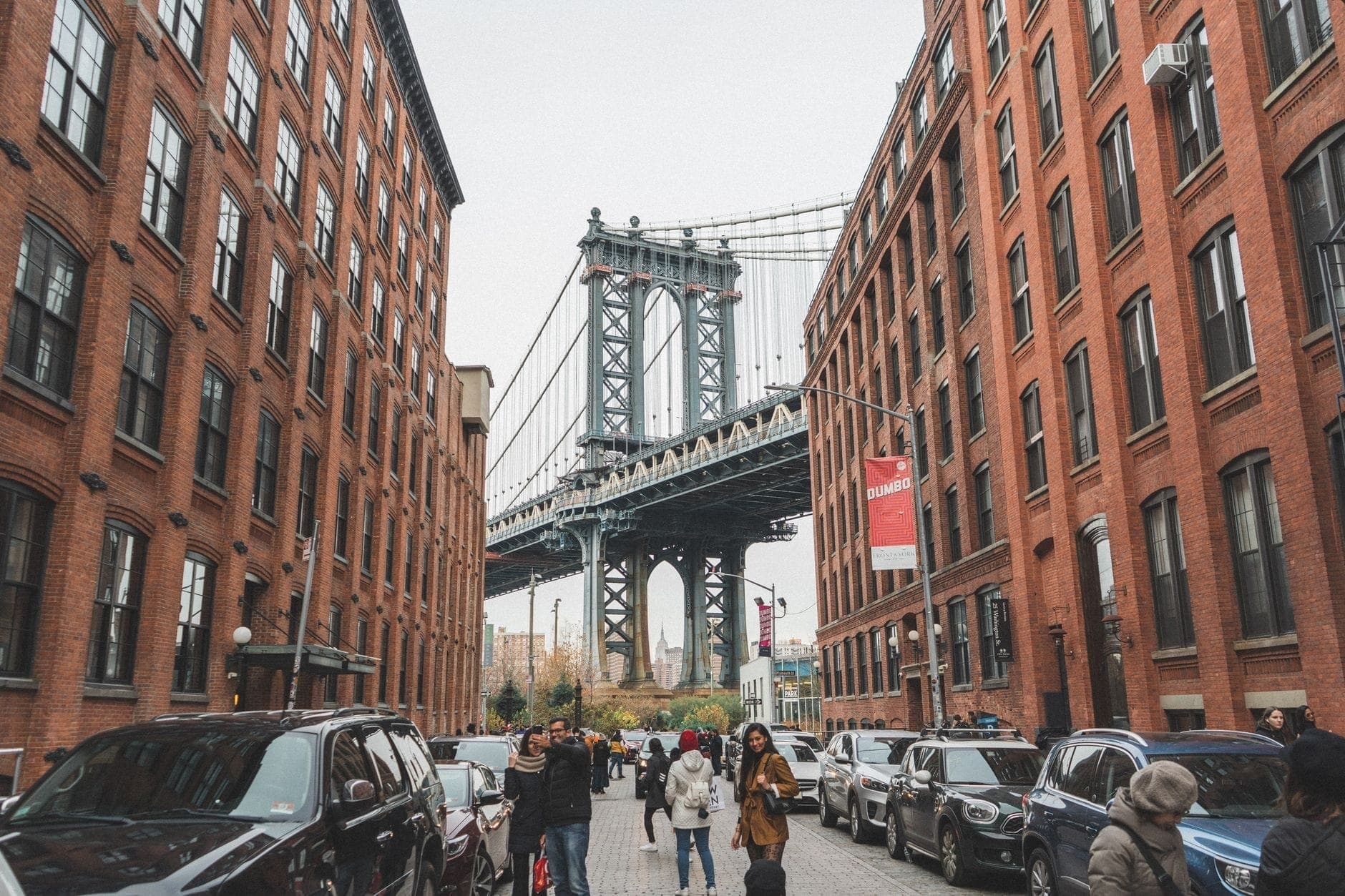วิวของ Manhattan Bridge ที่ถูกล้อมรอบด้วยอาคารอิฐสีแดงใน DUMBO บรูคลิน พร้อมผู้คนเดินอยู่บนถนนหินกรวด