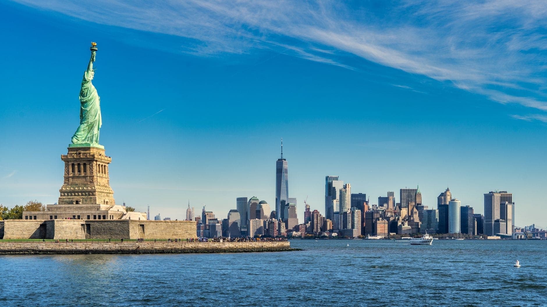 อนุสาวรีย์เทพีเสรีภาพในเบื้องหน้า พร้อมเส้นขอบฟ้าของ Lower Manhattan และ One World Trade Center ในวันที่แจ่มใสและแดดจัด