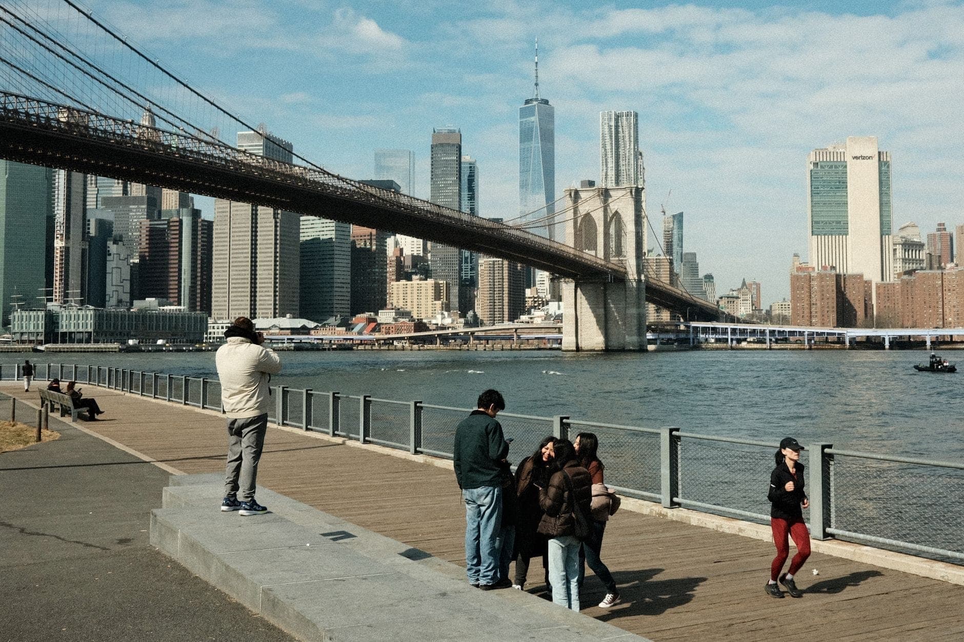 วิวของ Brooklyn Bridge พร้อมเส้นขอบฟ้าของ Manhattan มีครอบครัวและเด็กๆ เดินเล่นและถ่ายรูปตามแนวริมน้ำ East River