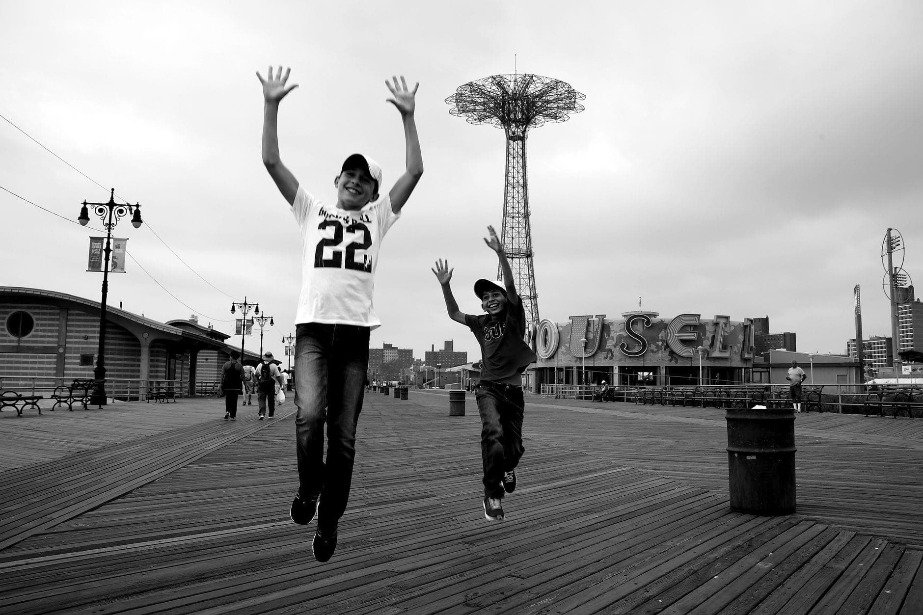 เด็กสองคนกระโดดบนทางเดินริมน้ำของ Coney Island โดยมี Parachute Jump และป้ายม้าหมุนอยู่เบื้องหลัง