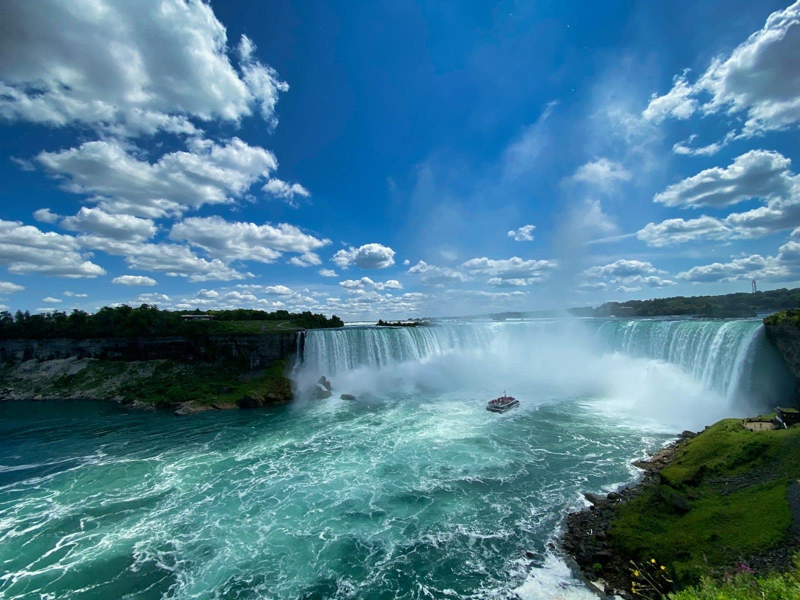 Vista amplia de las Cataratas del Niágara con agua turquesa, niebla que se eleva, barco turístico cerca de las cataratas, paisaje verde y cielo azul lleno de nubes.