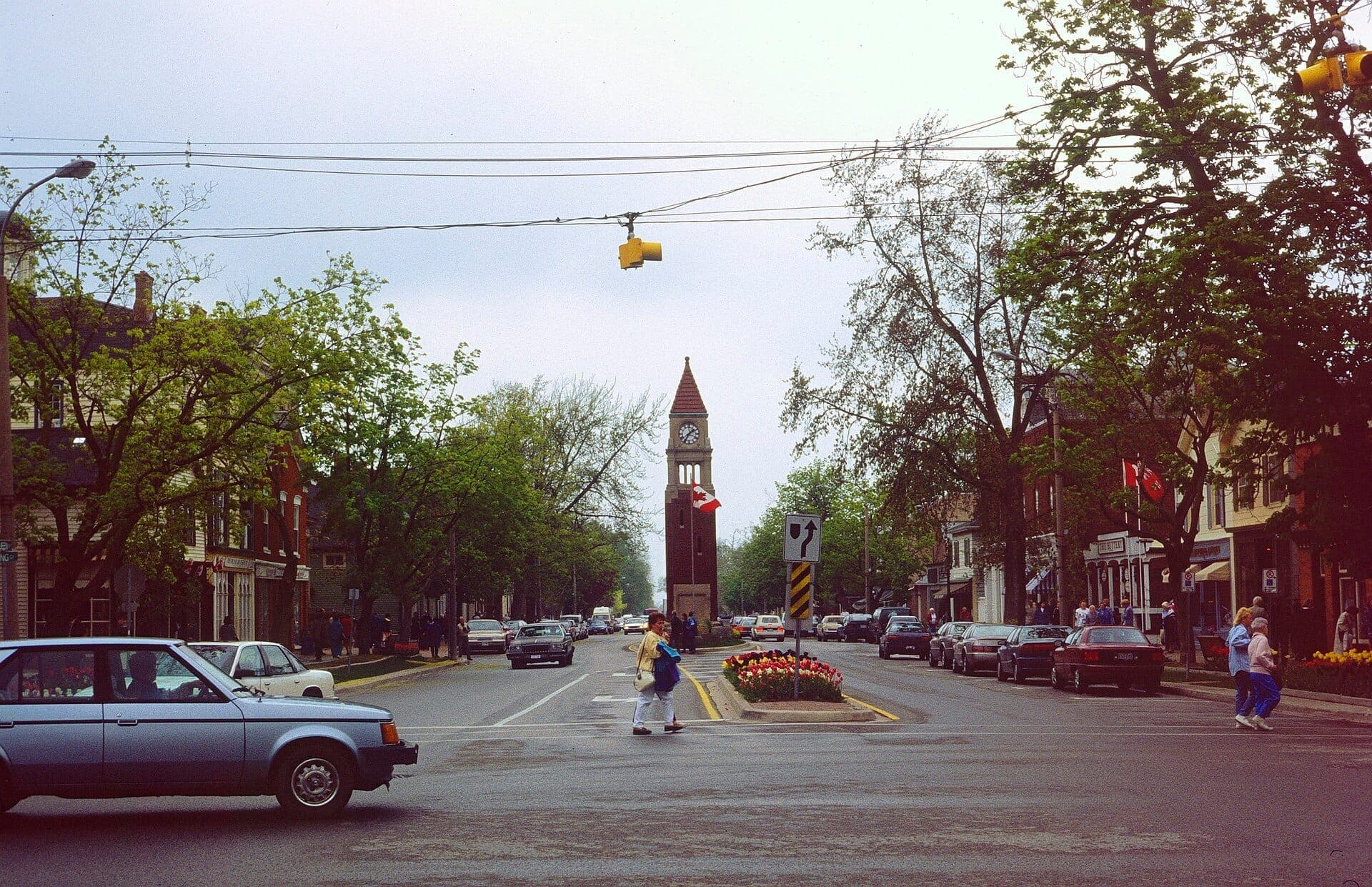 Calle principal histórica de Niagara-on-the-Lake con torre del reloj, autos antiguos, tiendas y peatones en un día nublado de primavera.