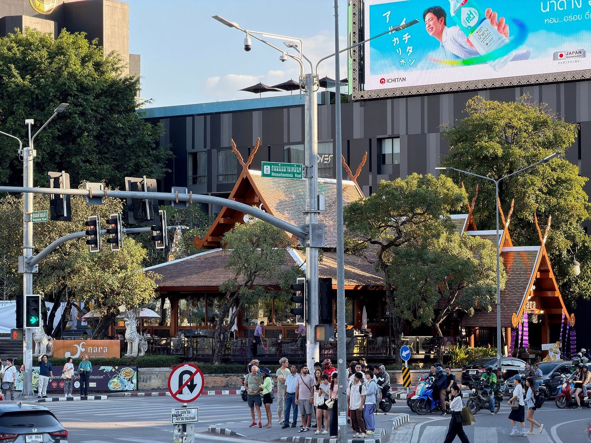 Belebte Kreuzung an der Nimmanhaemin Road in Chiang Mai mit modernen Gebäuden, traditioneller thailändischer Architektur, Ampeln und Gruppen von Menschen, die die Straße überqueren.