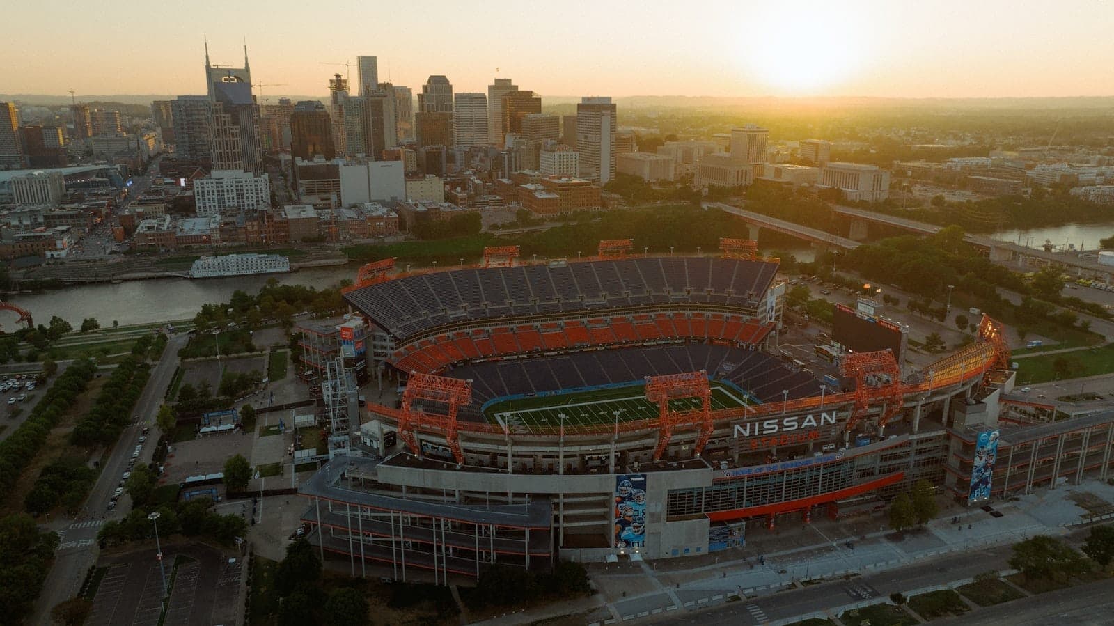 Aerial view of Nissan Stadium at sunset with downtown Nashville skyline in the background, nestled along the Cumberland River under a golden sky.