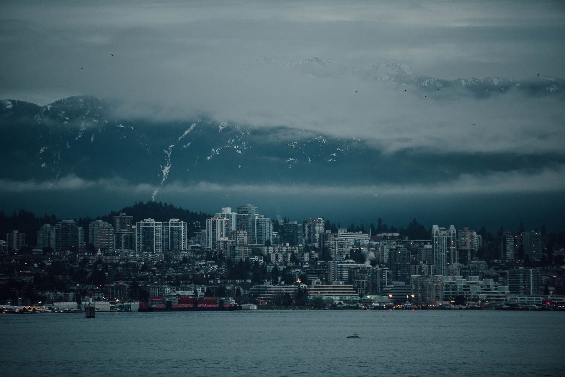 วิวเมือง North Vancouver กับฉากหลังเป็นภูเขาสูงตระหง่าน มีเมฆจับยอดเขาและ Burrard Inlet อยู่ข้างหน้า แสงบรรยากาศหม่นอมเทา