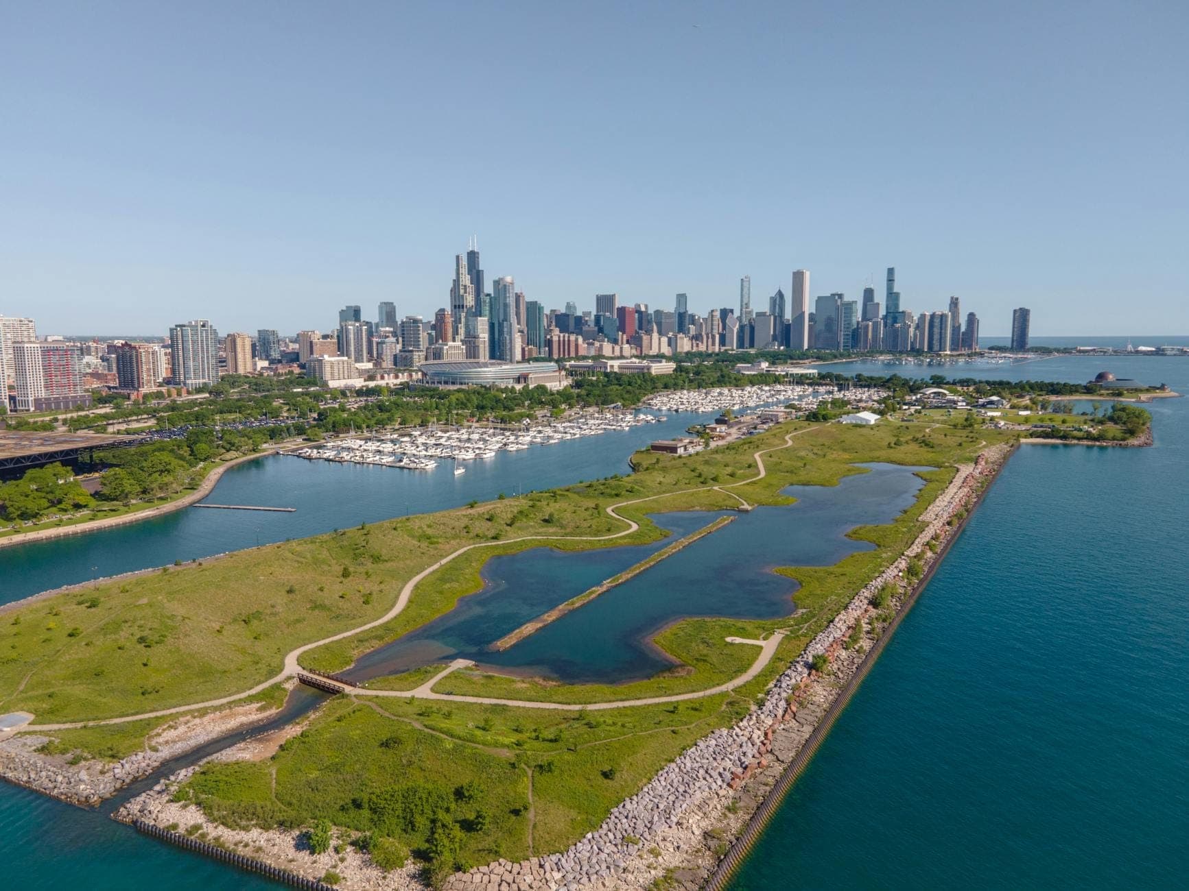A wide aerial view of Northerly Island Park with winding trails, restored prairie, Lake Michigan, and the Chicago skyline in the background on a clear day.