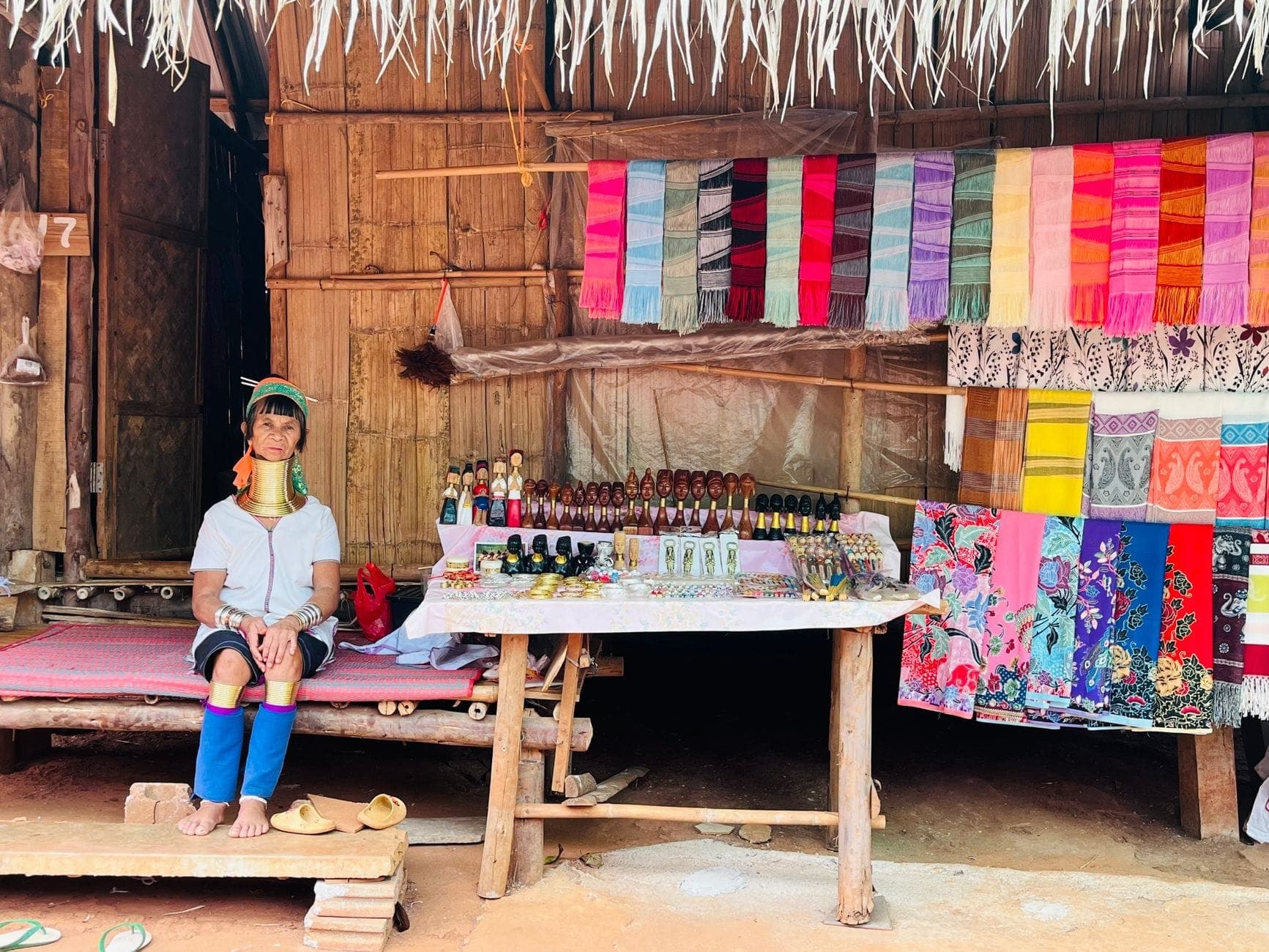 Una mujer con vestimenta tradicional de tribu de montaña y anillos de latón en el cuello sentada junto a una mesa de artesanías y textiles en un pueblo del norte de Tailandia.