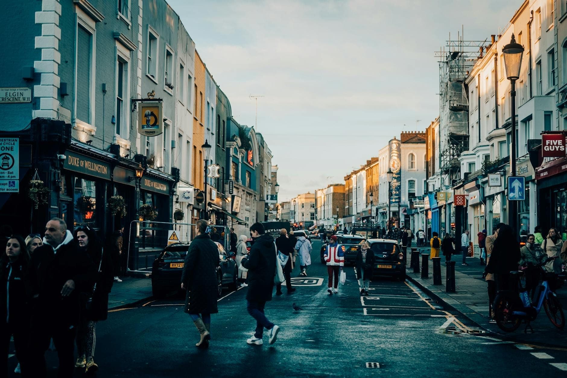 Calle concurrida en Notting Hill con personas cruzando, casas adosadas de colores, tiendas y arquitectura londinense característica, en un ambiente que evoca la ruta del desfile del carnaval.