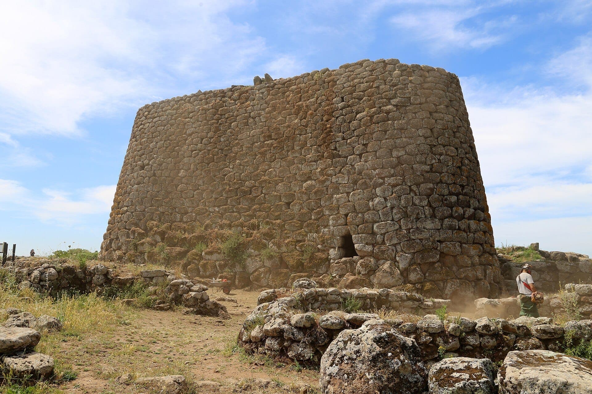 La enorme torre de piedra del Nuraghe Losa se eleva sobre ruinas dispersas y hierba silvestre bajo un cielo azul brillante en Cerdeña.